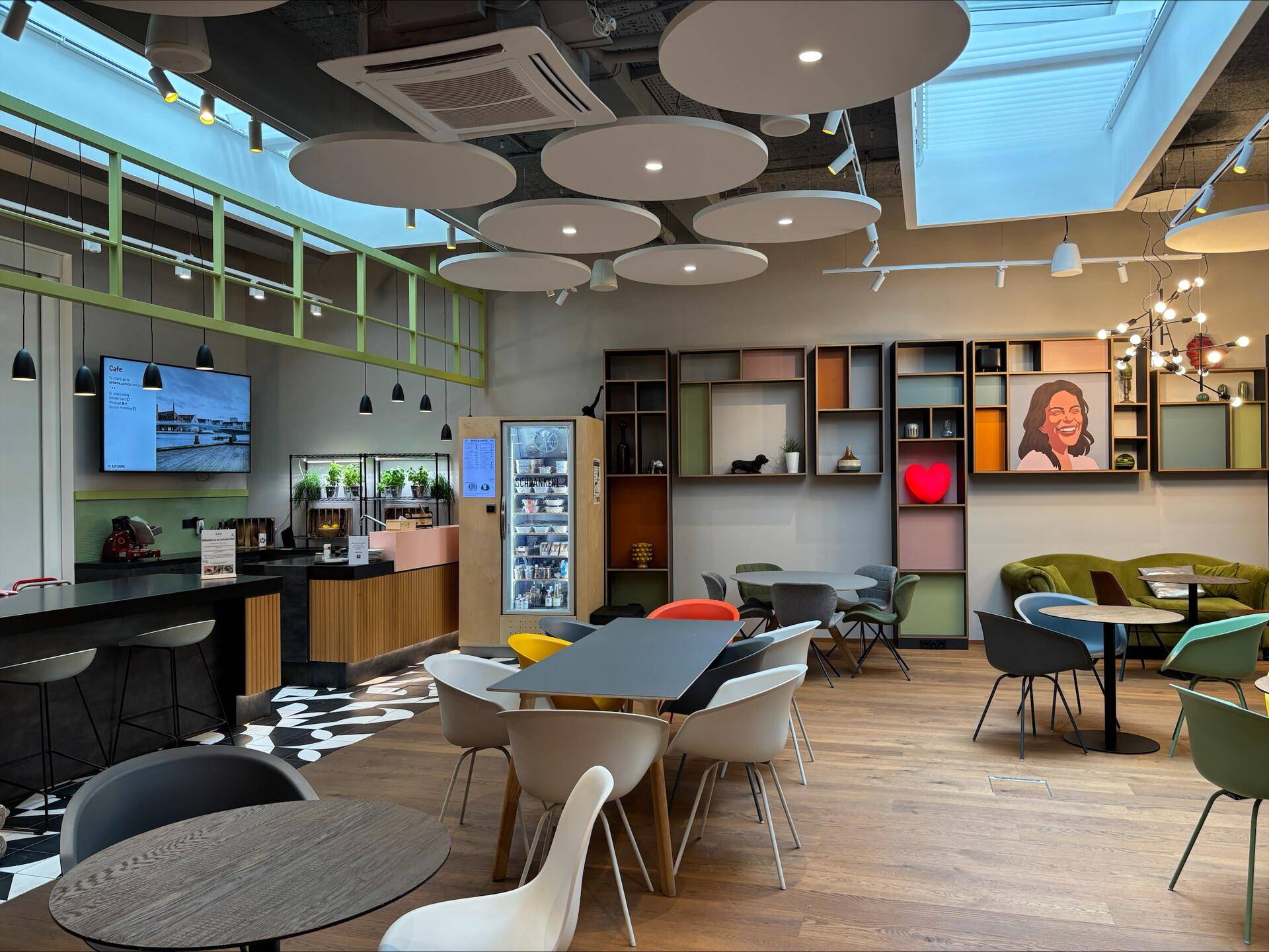 Heller Café-Bereich mit verschiedenen Tischen, Stühlen und einer Selbstbedienungsstation in einem modernen Ambiente.