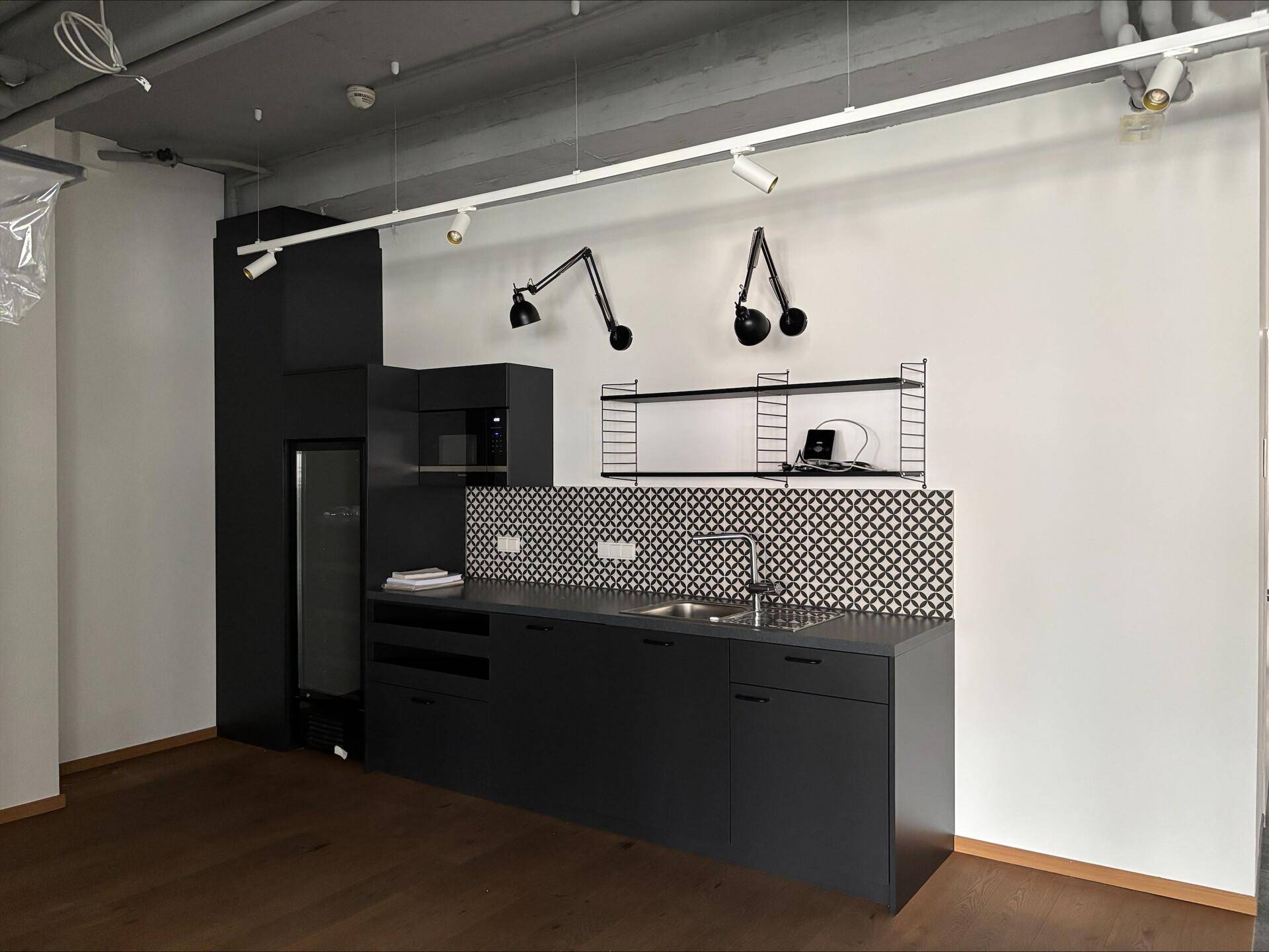 Moderne schwarze Küchenzeile mit Spüle, Kühlschrank und Wandregalen in einem funktionalen Büroraum.