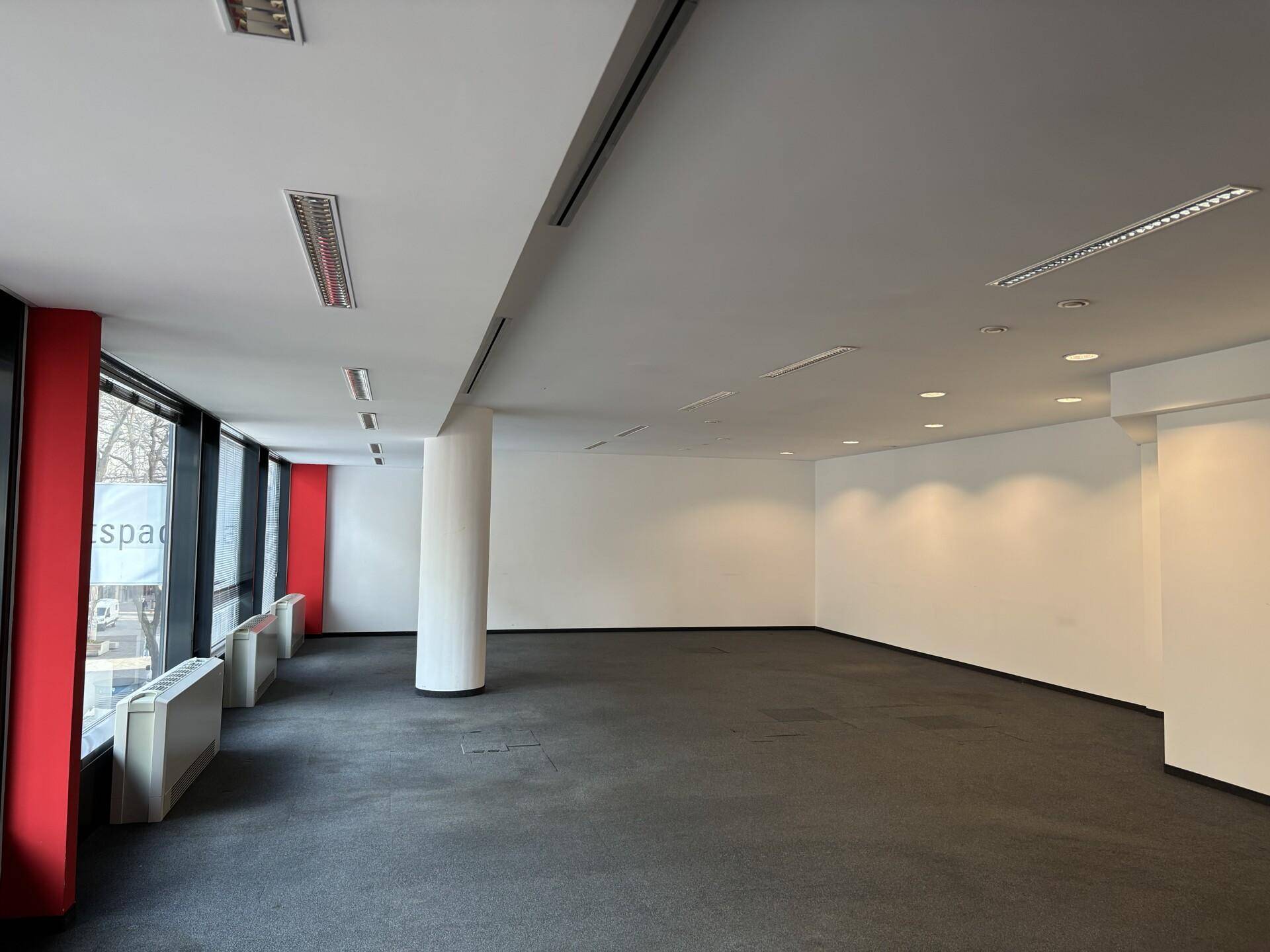 Geräumiger, leerer Büroraum mit Teppichboden, einer Säule und großen Fenstern, die viel Tageslicht hereinlassen.