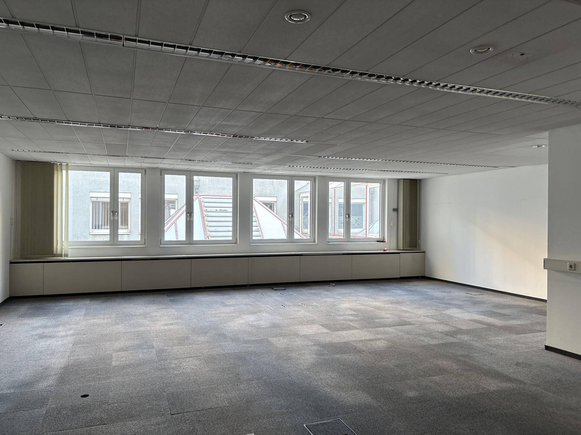 Büroraum mit Teppichboden, einer Reihe von Fenstern und niedrigen Einbauschränken entlang der Wand.