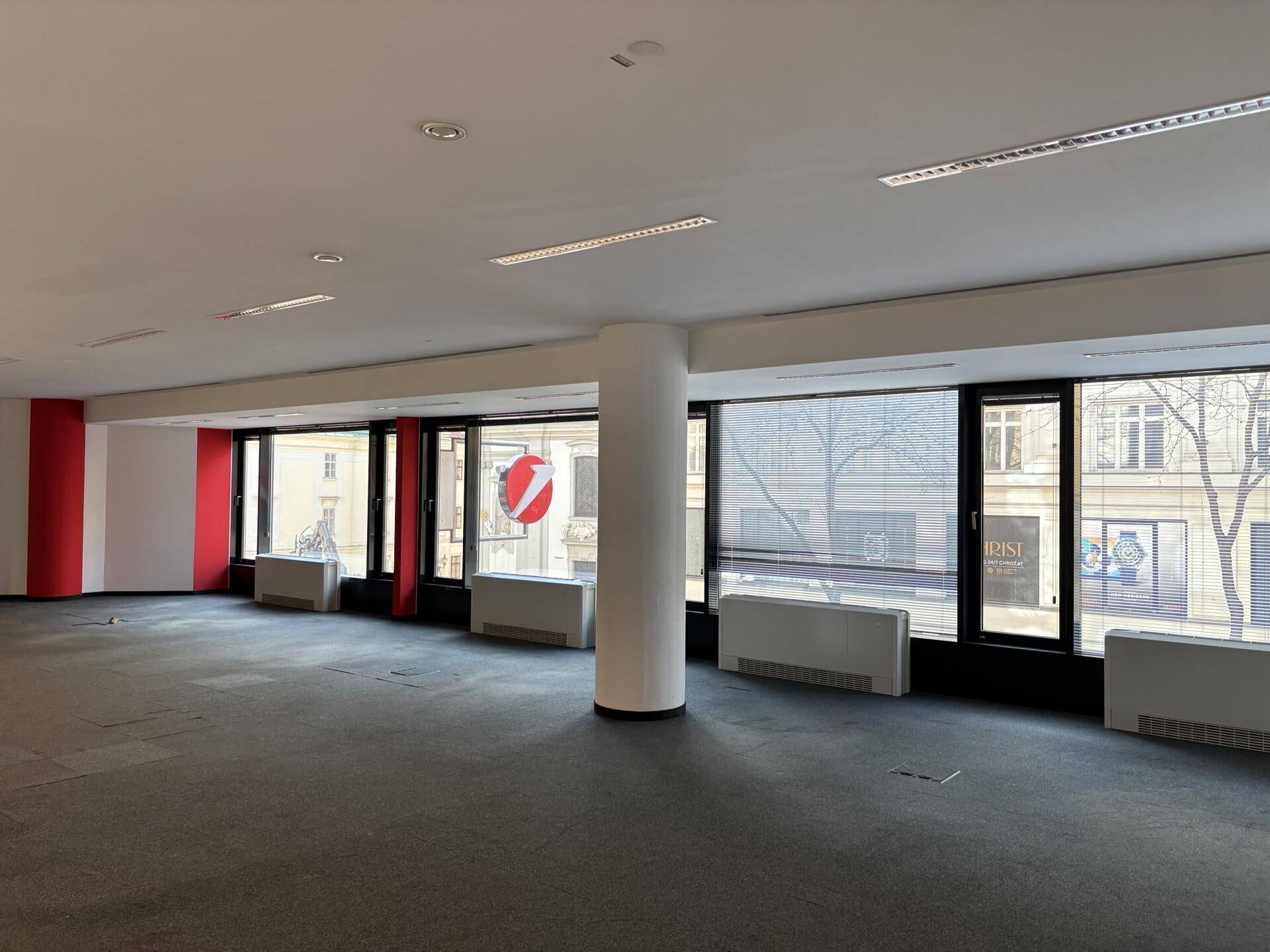 Heller Büroraum mit Teppichboden, einer zentralen Säule und großen Fenstern, die den Blick auf die Umgebung freigeben.