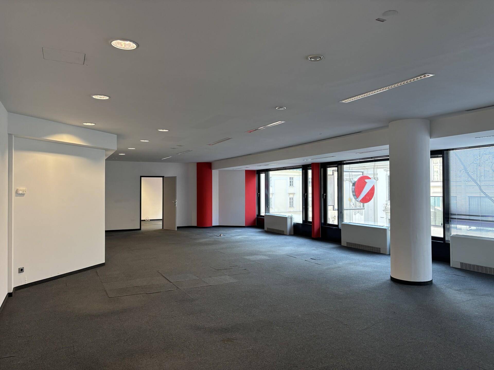 Großer, leerer Büroraum mit Teppichboden, weißen Wänden und einer Fensterfront mit Blick auf die Stadt.