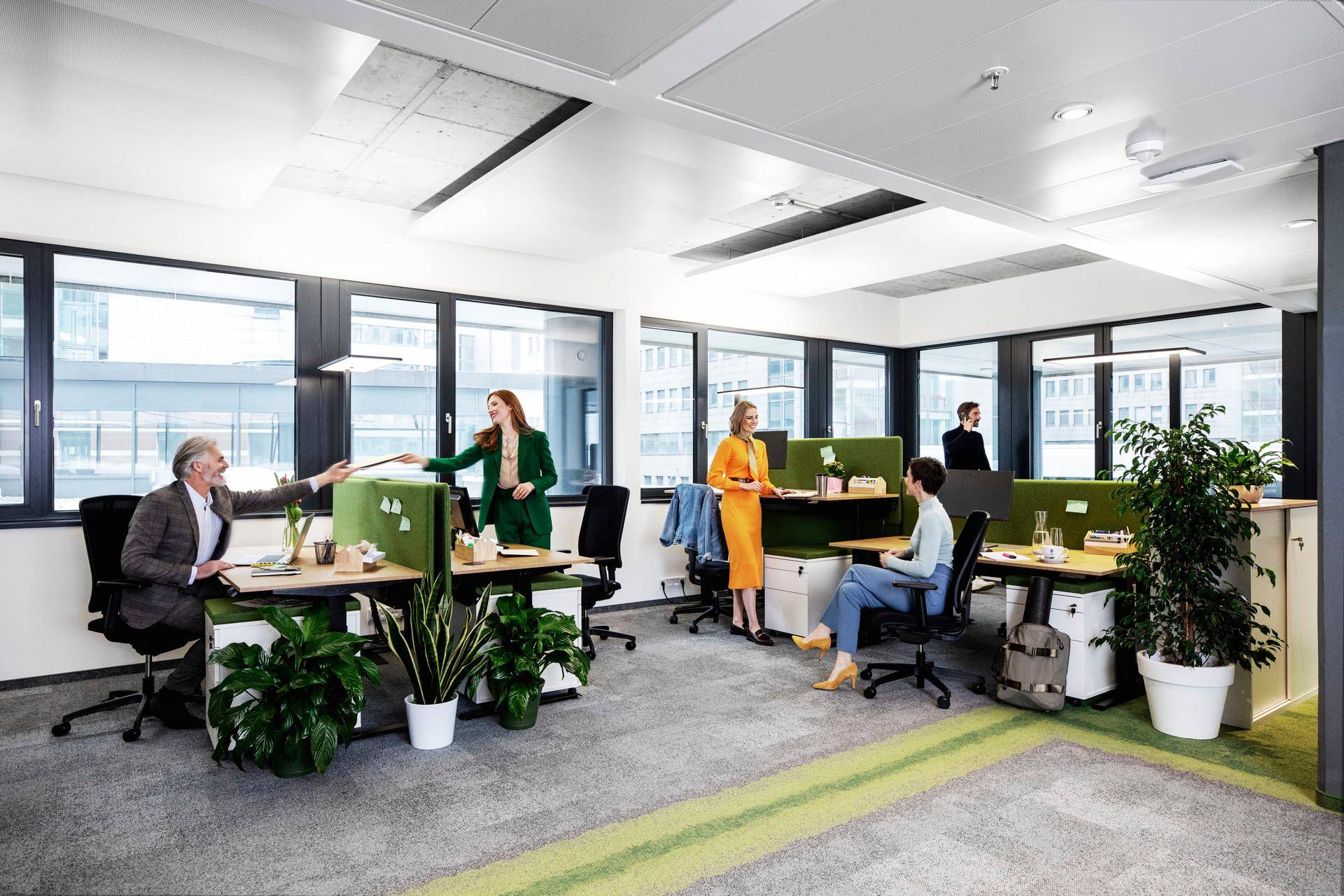 Helles Großraumbüro mit modernen Arbeitsplätzen, Pflanzen und großen Fenstern.