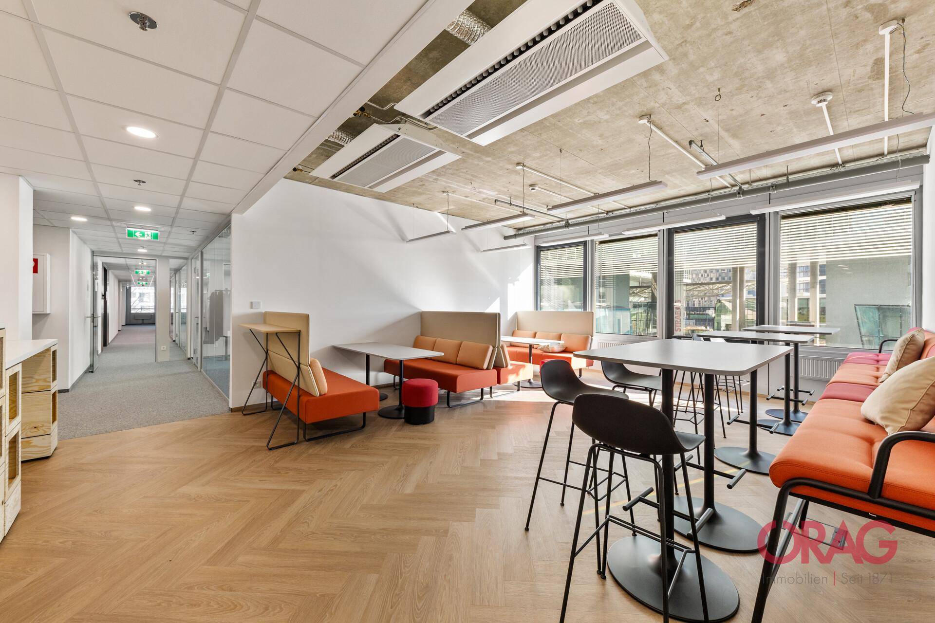 Moderner Pausenraum mit orangefarbenen Sitzgelegenheiten, Holzboden in Fischgrätmuster und industrieller Decke.