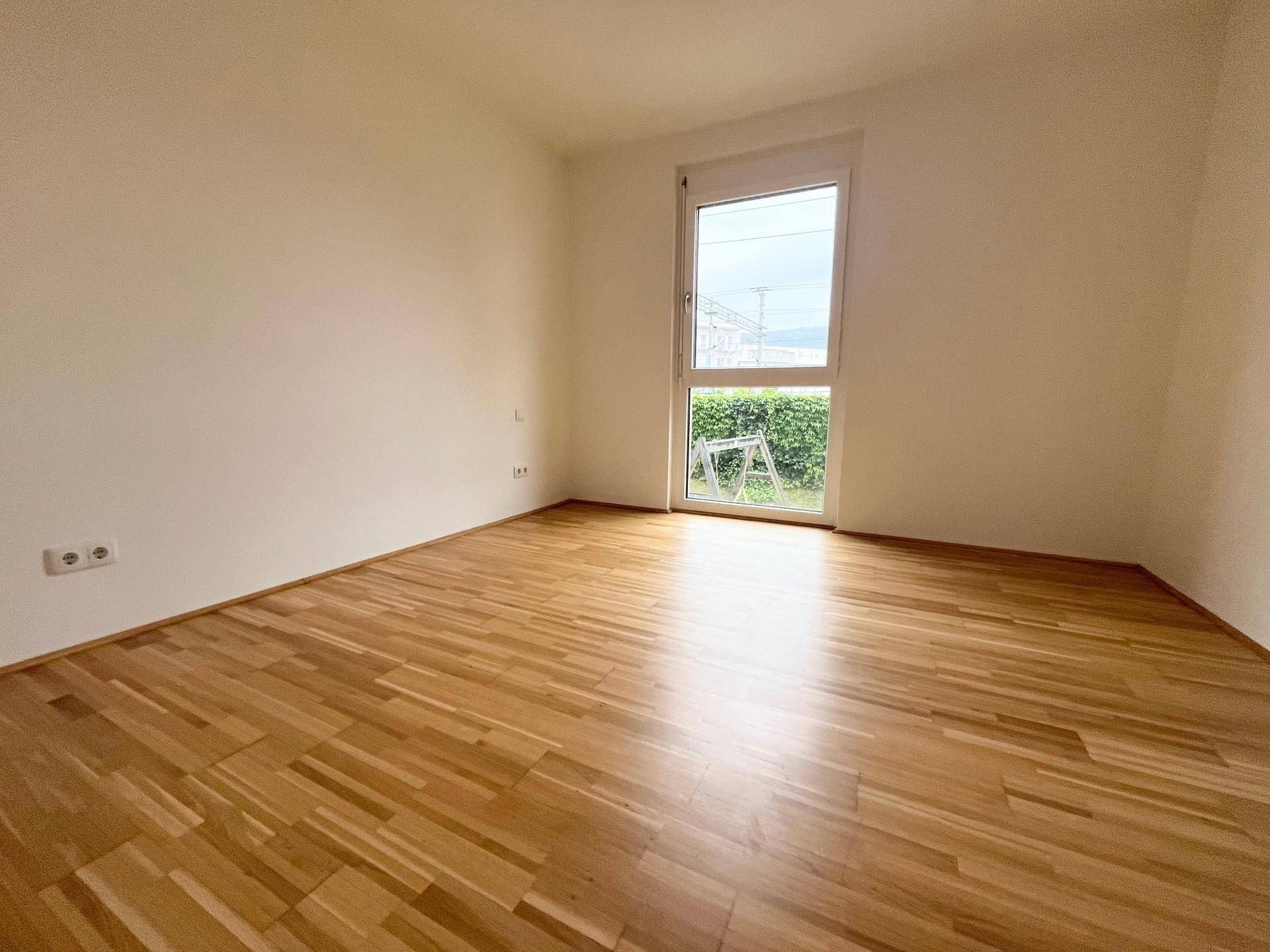 Helles Schlafzimmer mit Parkettboden und großem Fenster mit Blick ins Grüne.