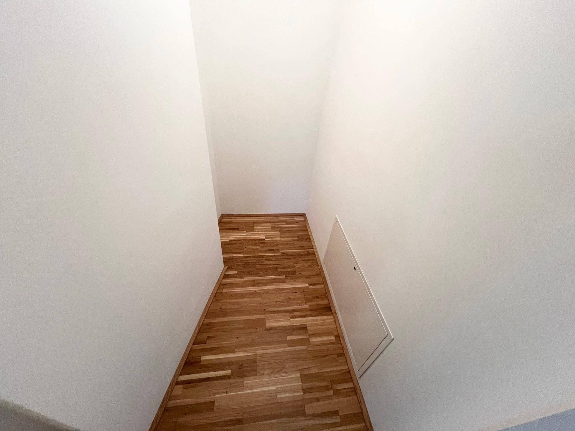 Schmaler Flur mit Holzparkett und weiß gestrichenen Wänden, der zu einem weiteren Bereich führt.