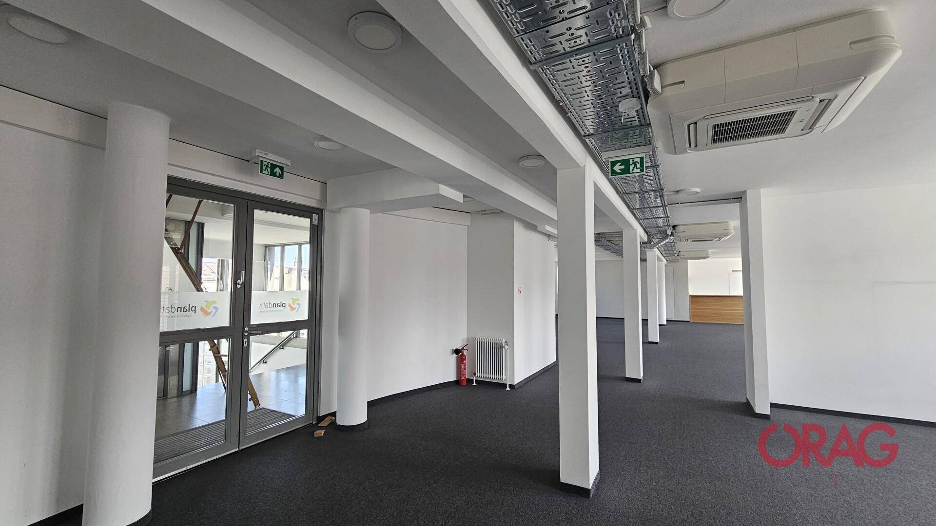 Leerer Büroraum mit Teppichboden, Säulen und sichtbaren Deckeninstallationen, Blick zum Treppenhaus.