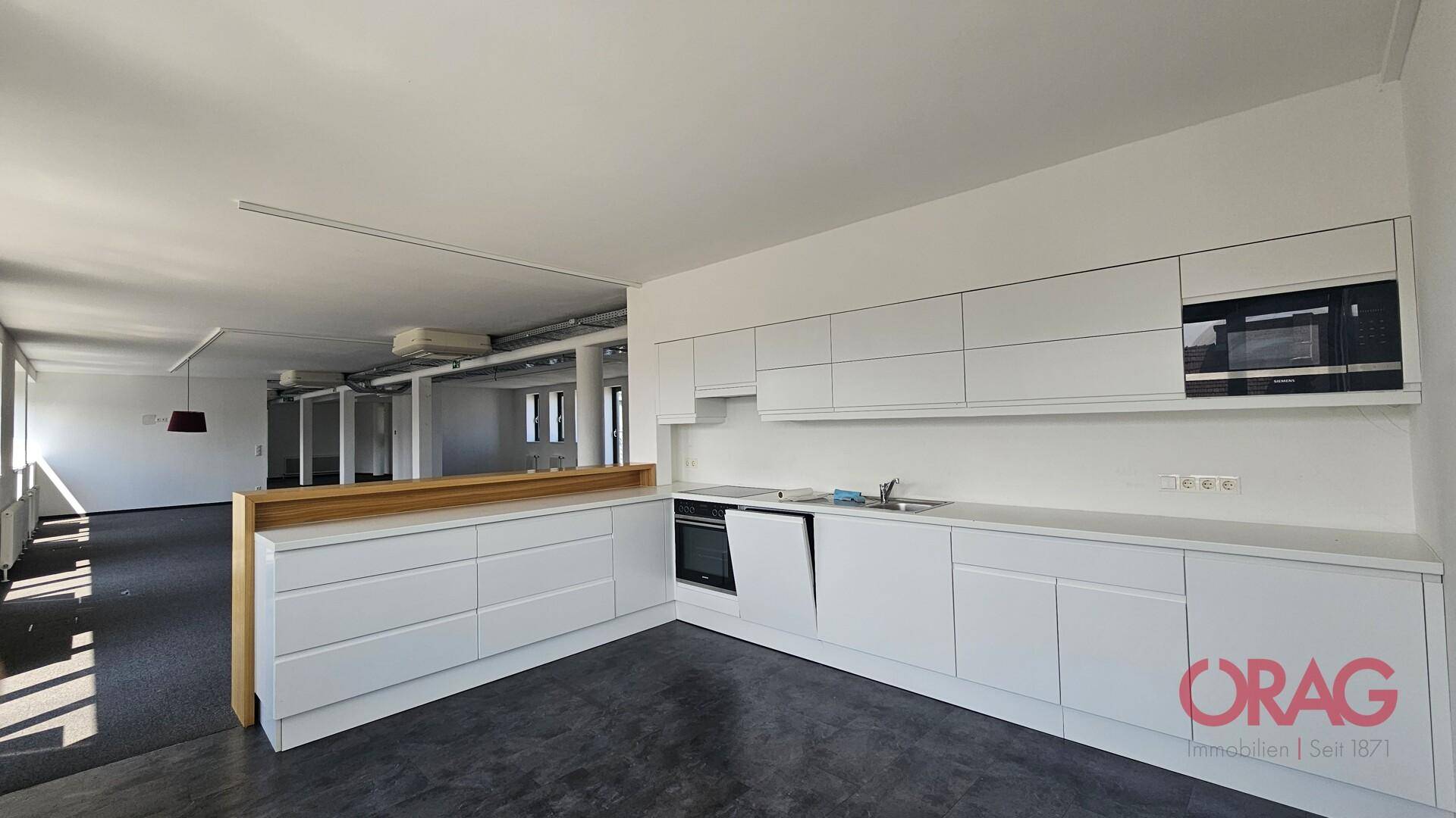 Moderne weiße Einbauküche mit integrierten Geräten und Holztheke in einem offenen Bürobereich.