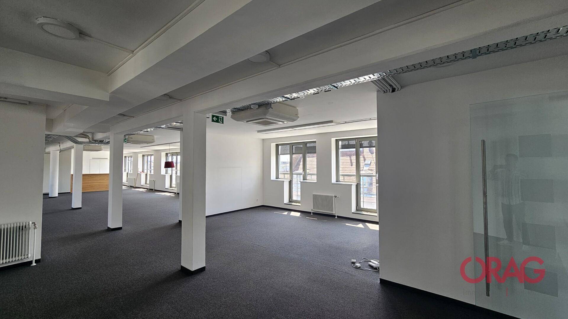 Großer, leerer Bürobereich mit Teppichboden, Säulen und Fenstern, die viel Tageslicht hereinlassen.