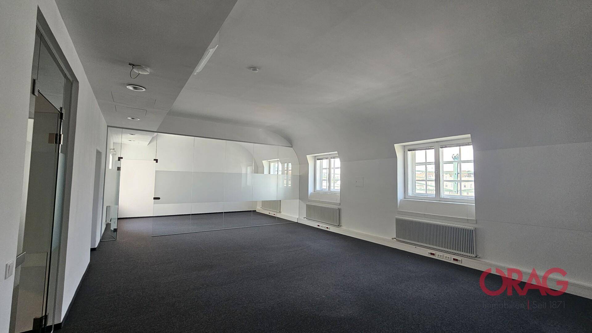 Geräumiger Büroraum mit Teppichboden, schrägen Decken und Fenstern, die eine einzigartige Atmosphäre schaffen.