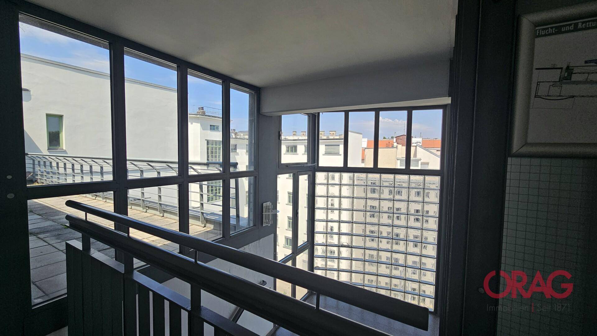 Helles Treppenhaus mit großen Fenstern, die einen weiten Blick auf die umliegenden Gebäude bieten.