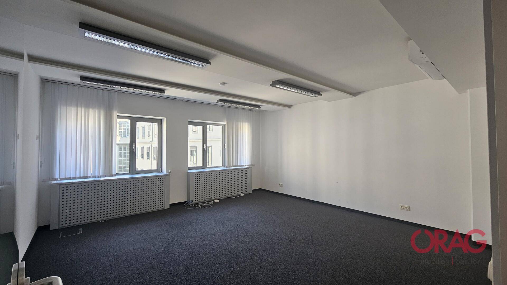 Helles Büro mit Teppichboden, großen Fenstern und vertikalen Jalousien, das viel Tageslicht bietet.