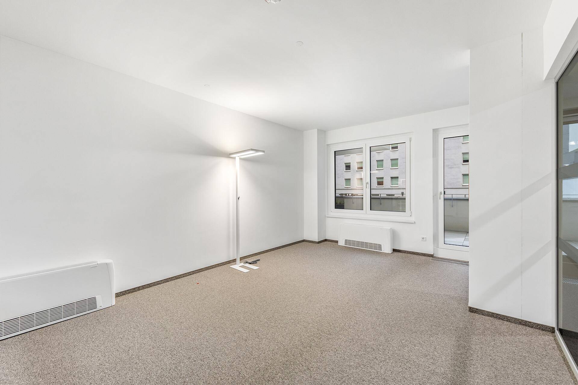 Leerer Büroraum mit Teppichboden, weiß gestrichenen Wänden und einem Fenster mit Blick auf andere Gebäude.
