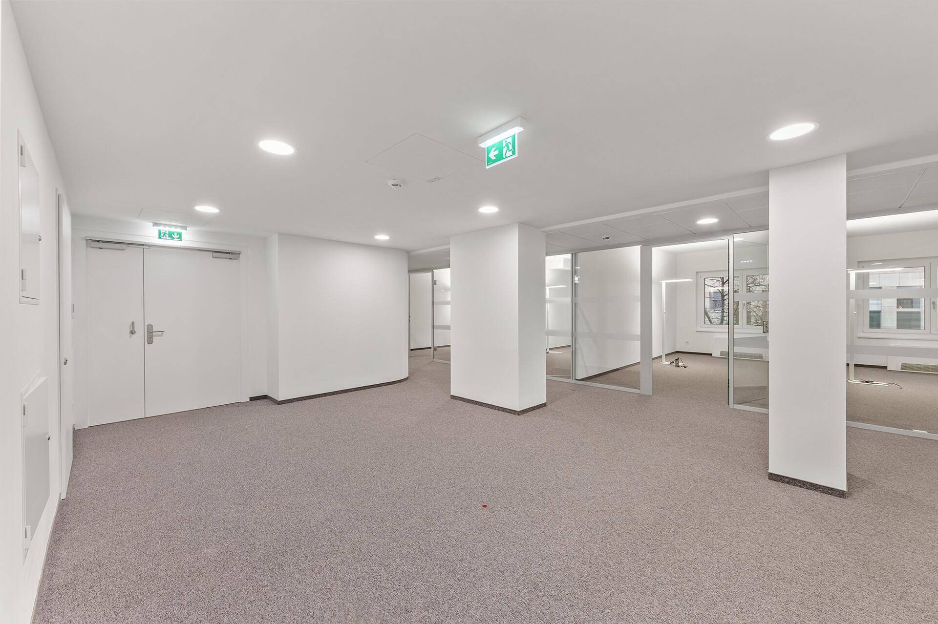 Moderner Büroraum mit Teppichboden, weißen Wänden und integrierten Deckenleuchten.