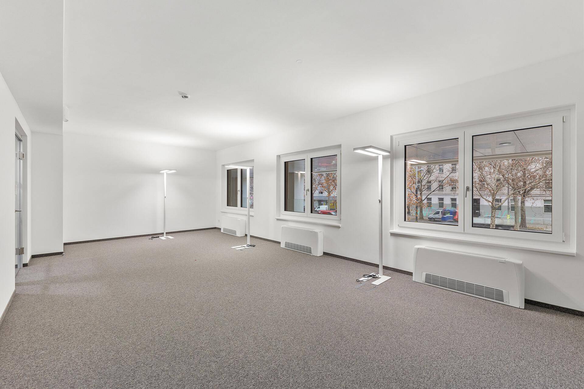 Geräumiges Büro mit Teppichboden, großen Fenstern und modernen Stehleuchten, bietet viel Tageslicht und Stadtblick.