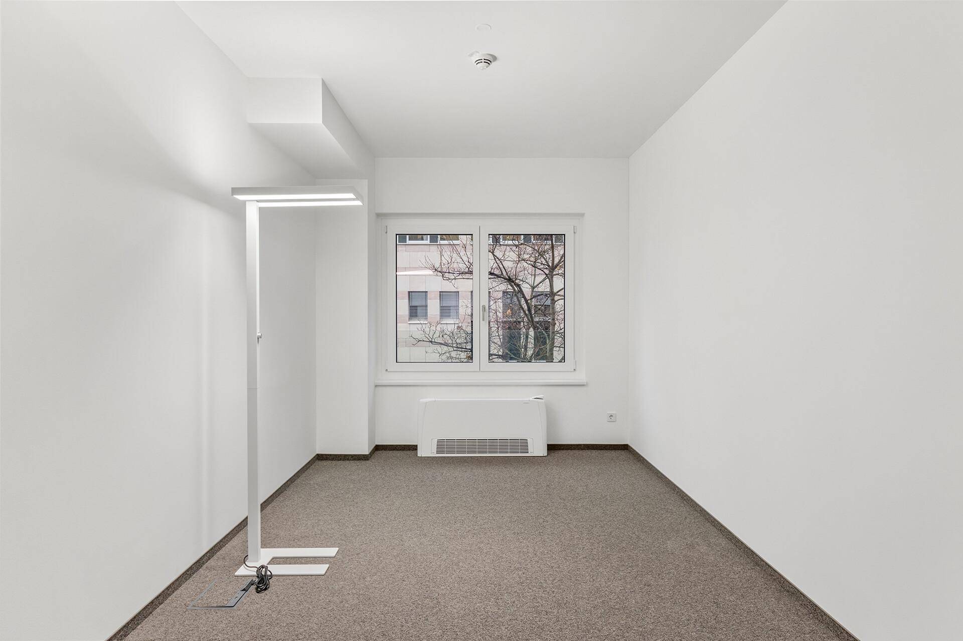 Kleines, helles Büro mit Teppichboden, einem Fenster und moderner Stehleuchte, ideal für Einzelarbeit.
