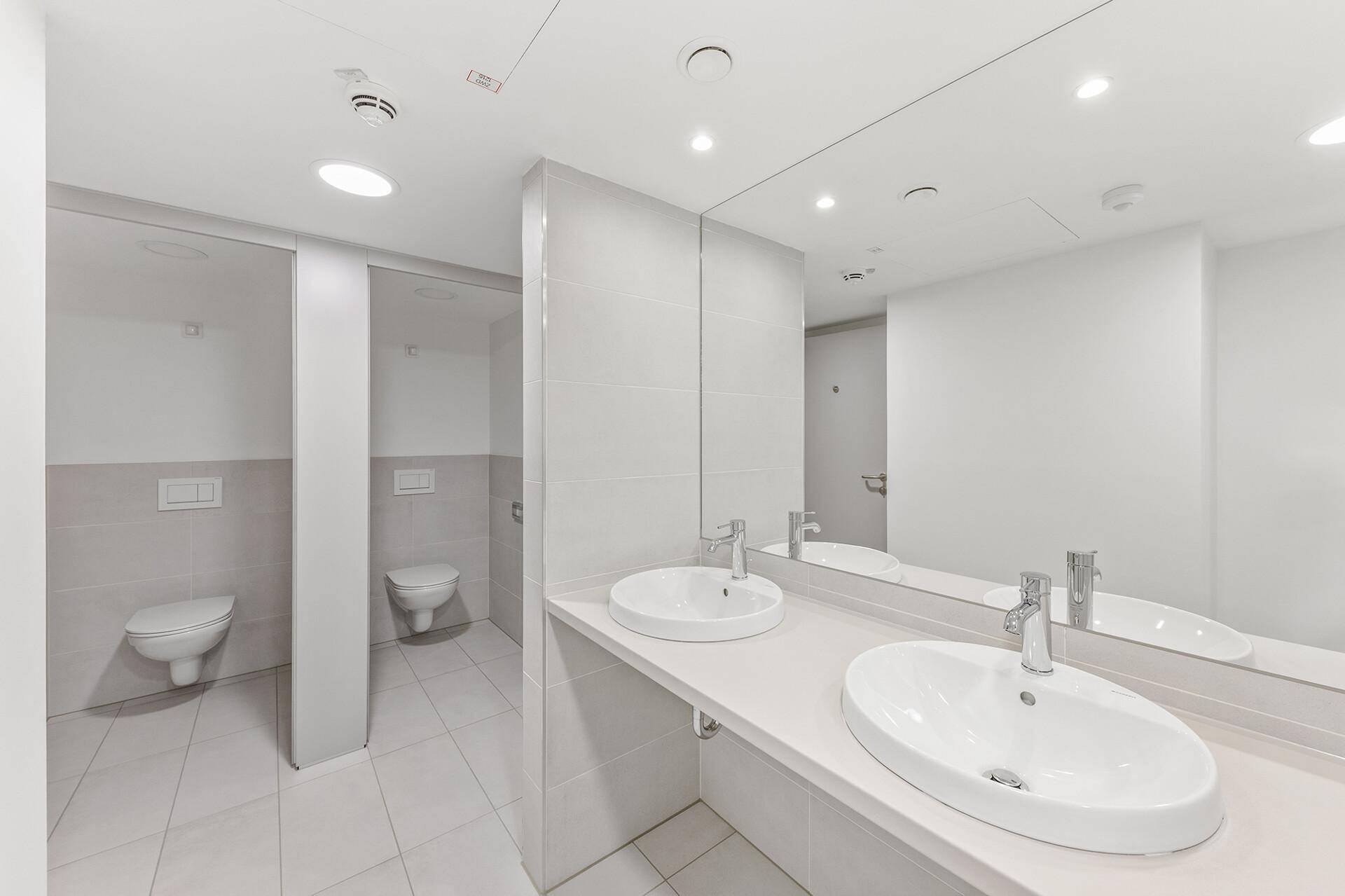 Moderne Toilettenanlage mit mehreren Kabinen, Doppelwaschbecken und großen Spiegeln, hell gefliest und sauber.