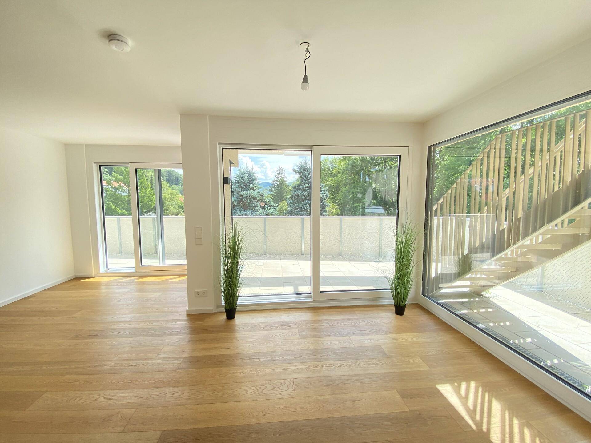 Geräumiger, leerer Wohnbereich mit hellen Holzböden und großen Fensterfronten mit Blick ins Grüne.