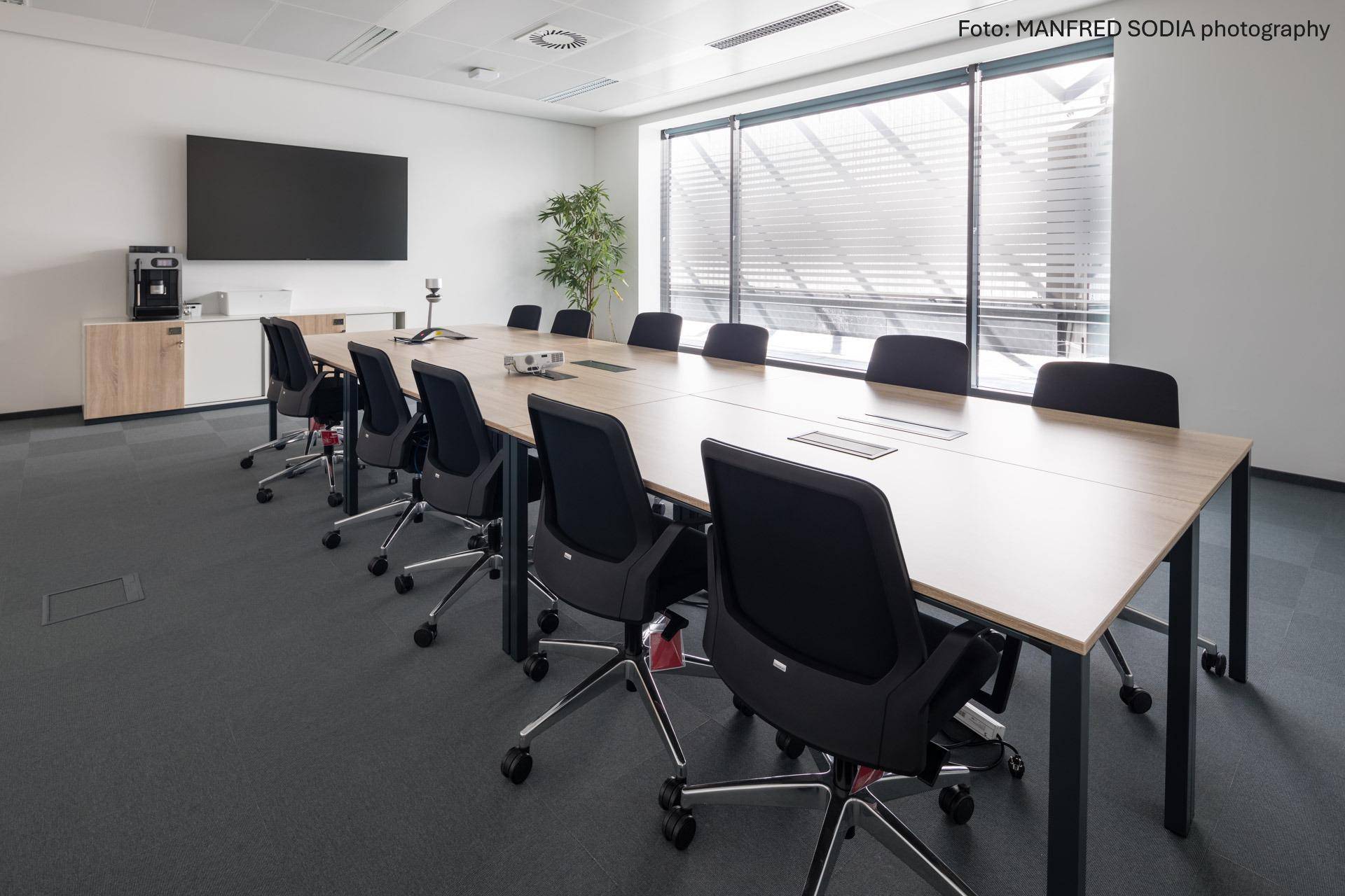 Moderner Konferenzraum mit großem Tisch, bequemen Stühlen und einem Wandbildschirm für Präsentationen.