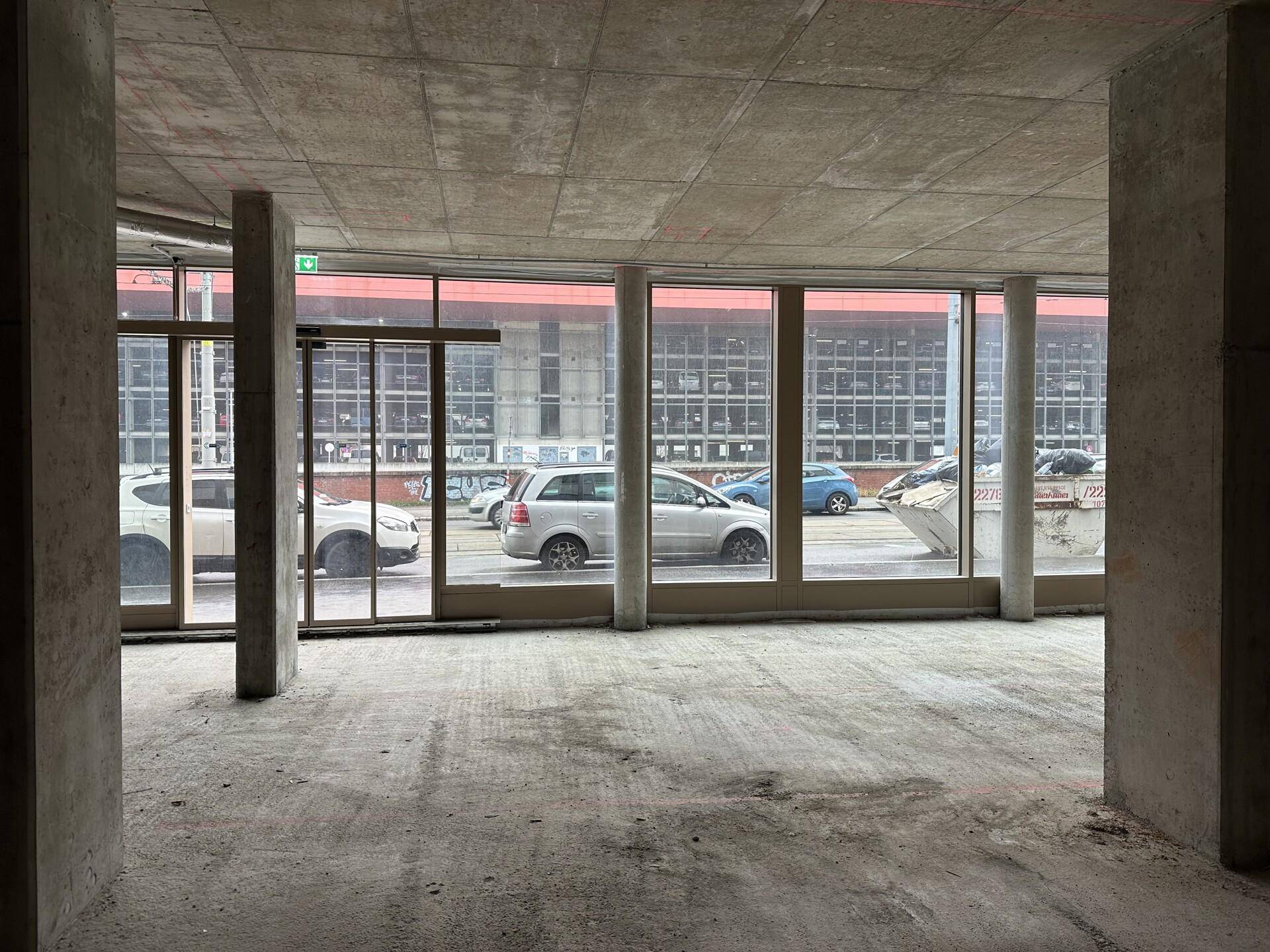Rohbau-Innenraum mit großen Fenstern zur Straße und Blick auf parkende Autos.