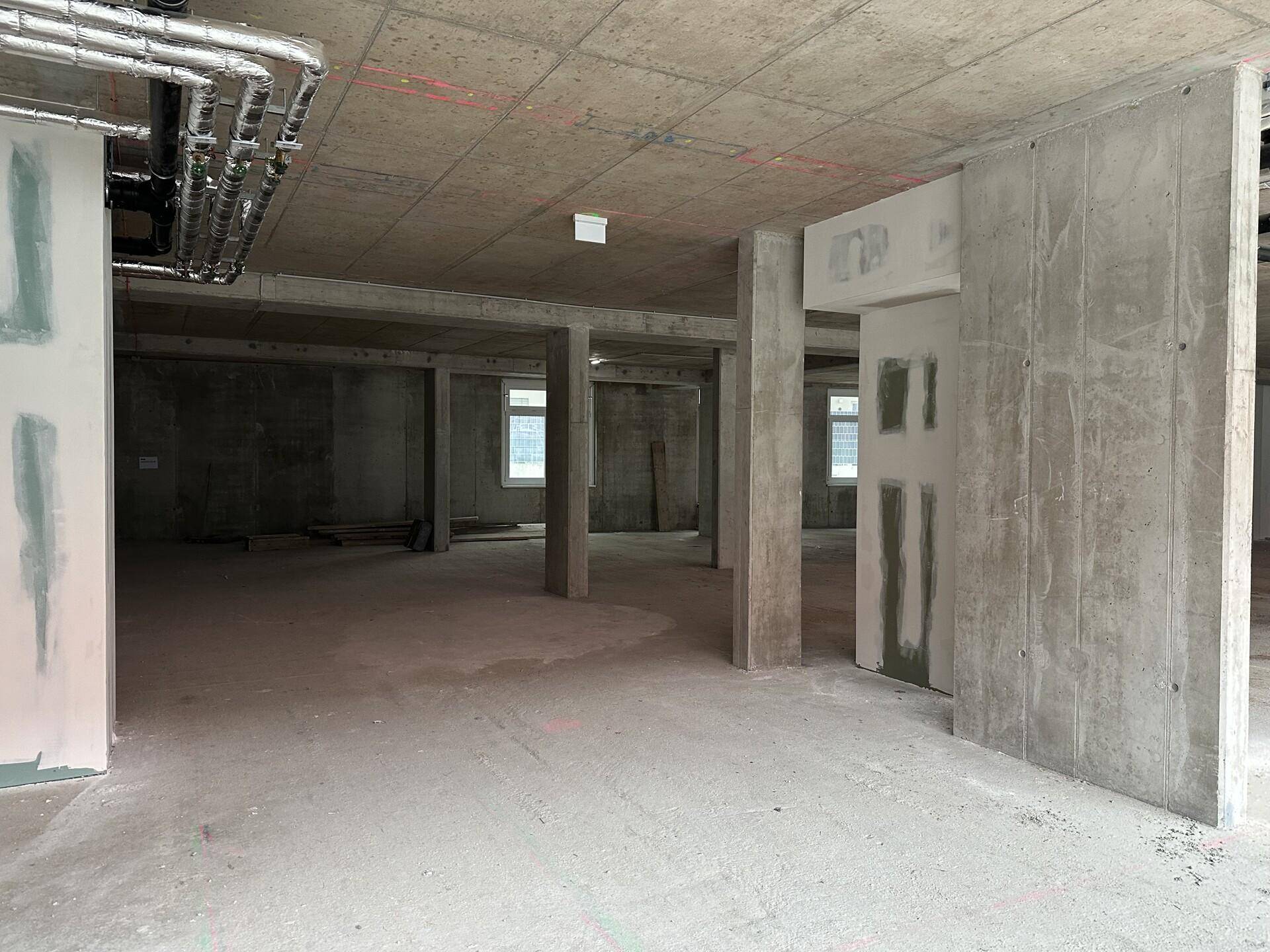 Rohbau-Innenraum mit sichtbaren Installationen und kleinen Fenstern im hinteren Bereich.