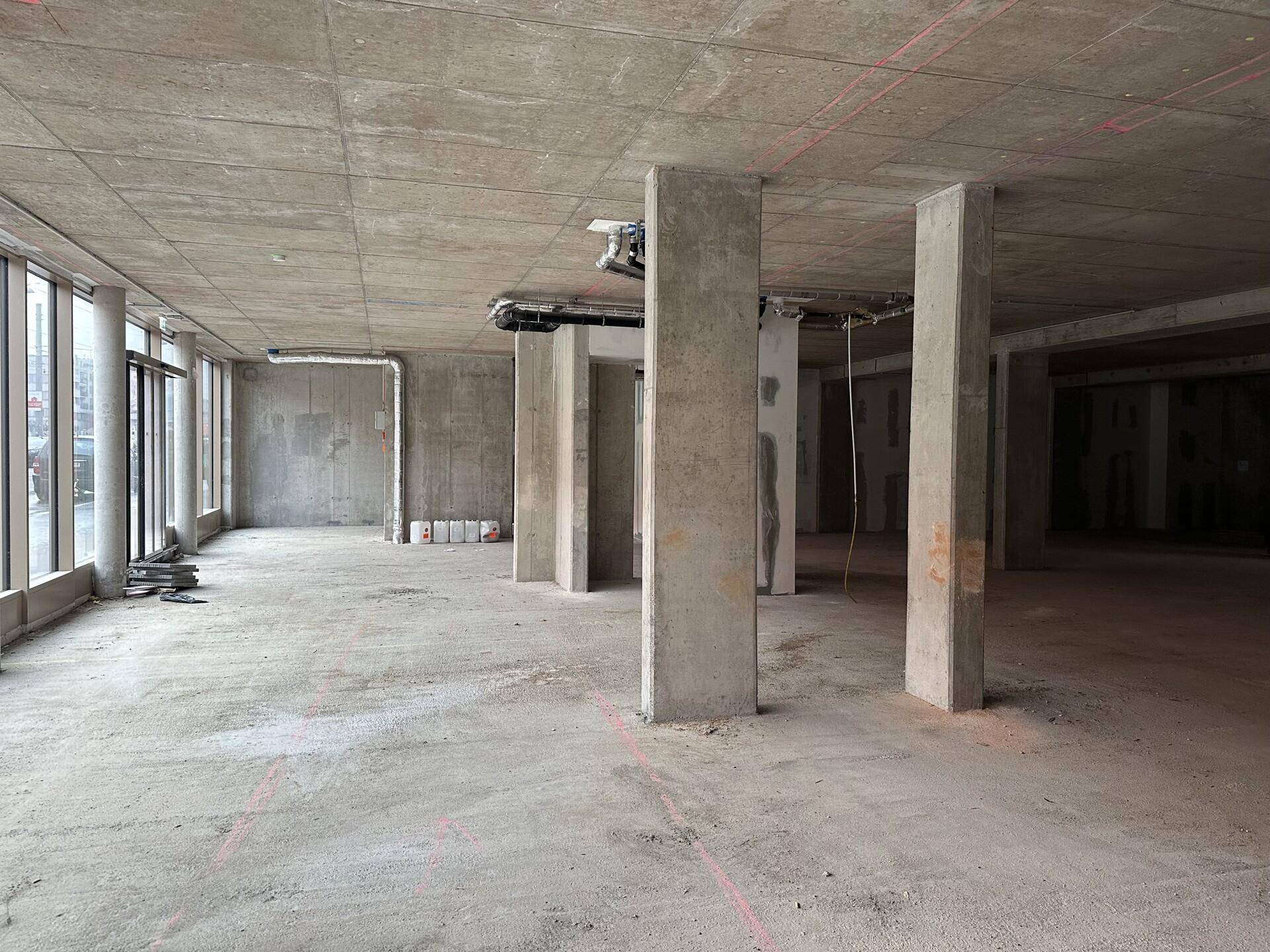 Offener Rohbau-Bereich mit Betonstützen und großen Fenstern, die viel Tageslicht hereinlassen.