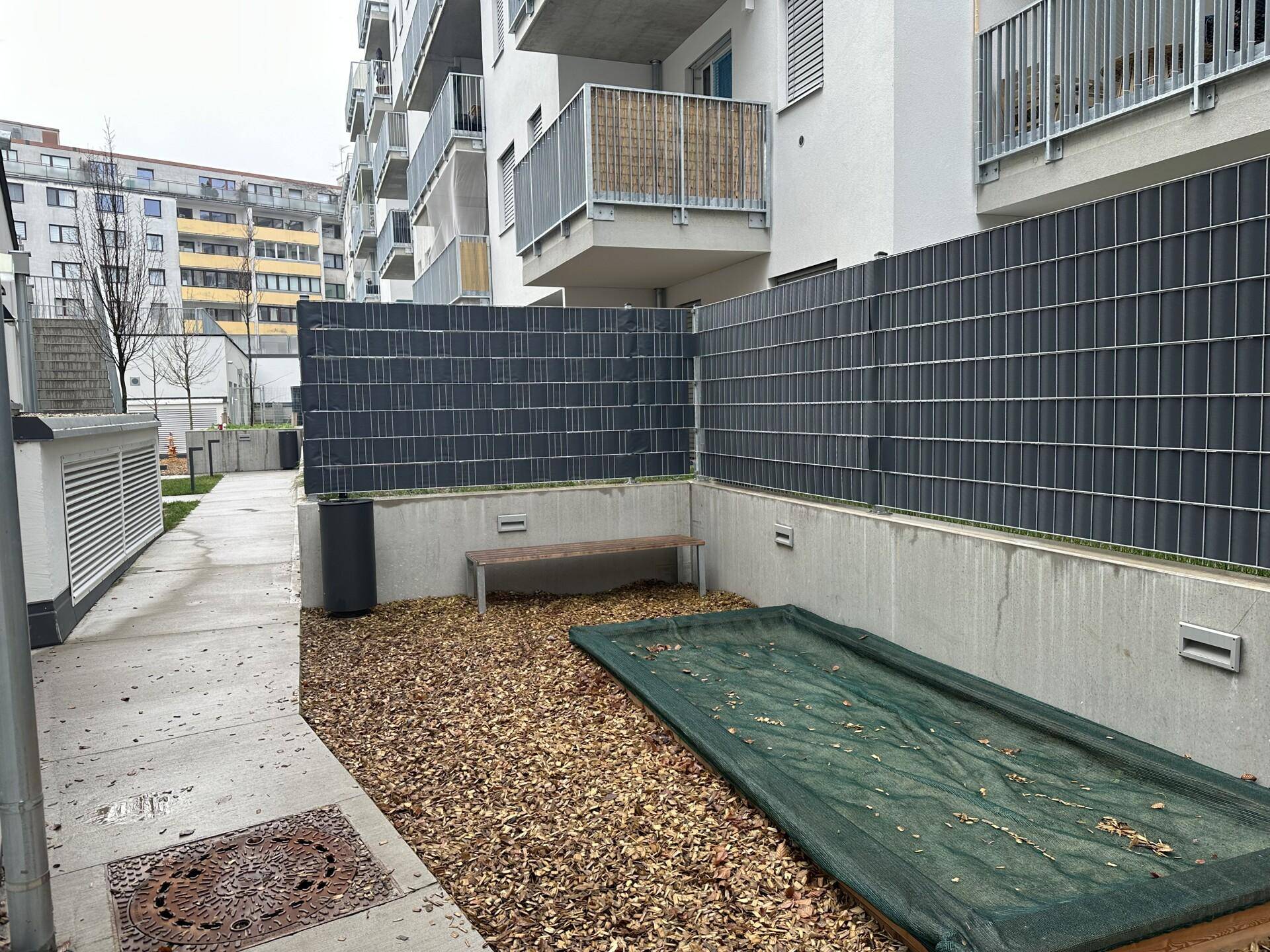 Kleiner, umzäunter Gartenbereich mit einer Bank und Laub, angrenzend an ein Wohngebäude.