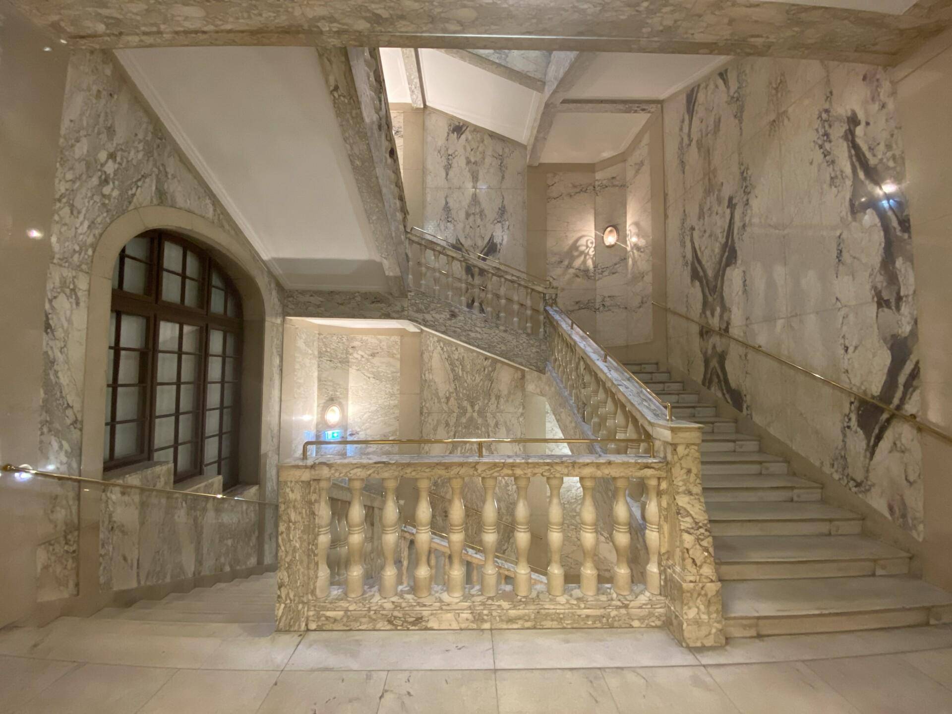 Elegantes Treppenhaus mit aufwendigen Marmorverkleidungen, kunstvollen Balustraden und einem großen Bogenfenster.