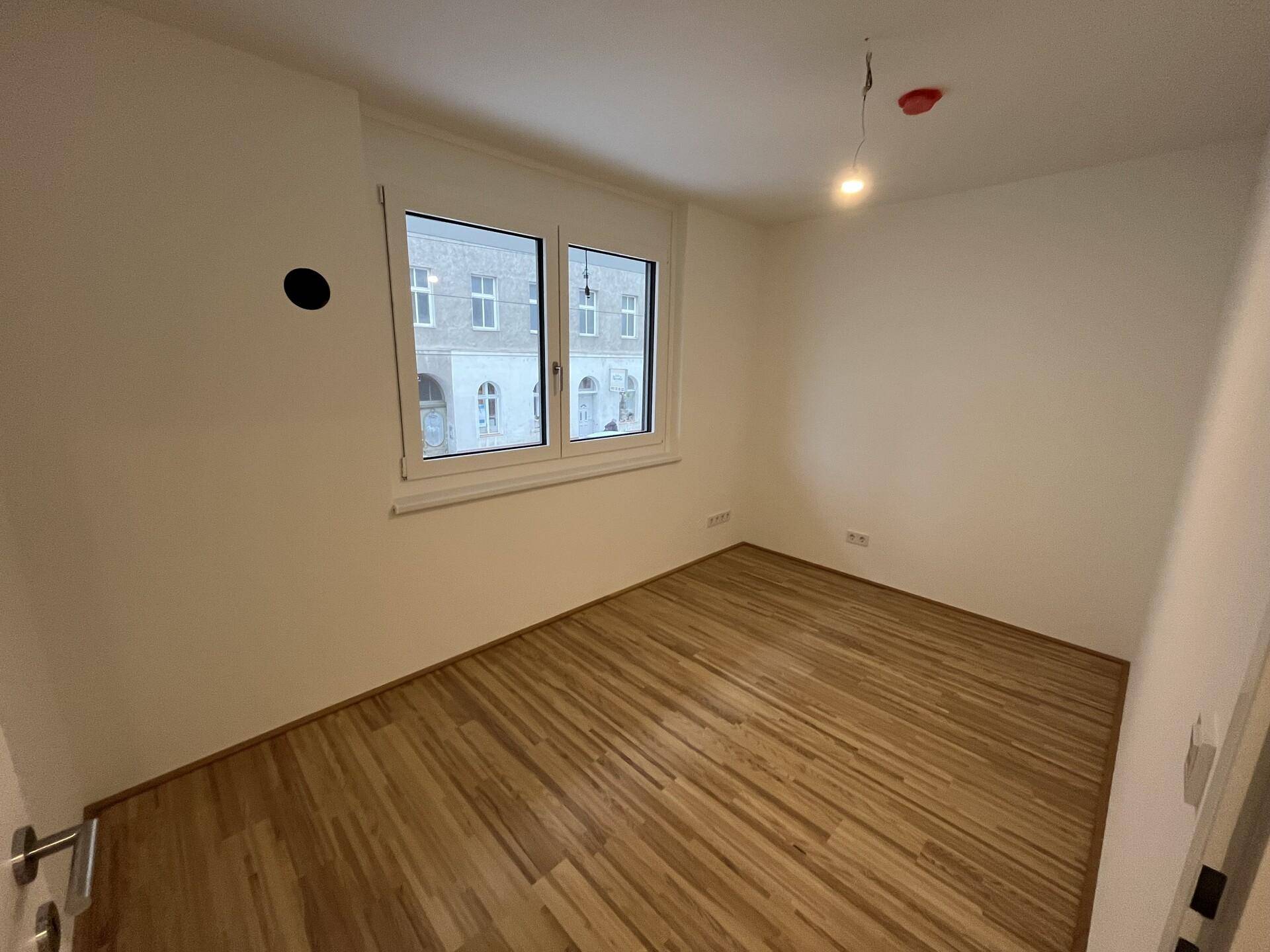 Leeres Zimmer mit Parkettboden und Fenster, das viel Tageslicht hereinlässt.