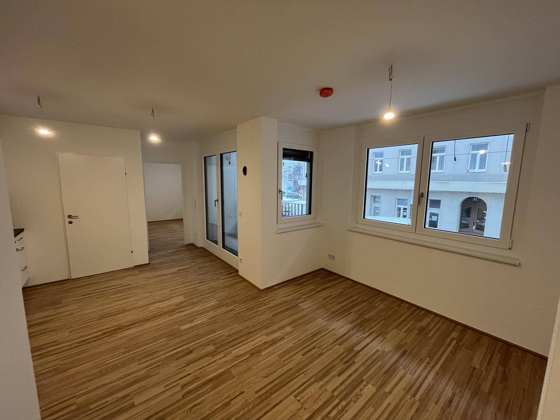 Geräumiges Wohnzimmer mit hellem Holzfußboden, großen Fenstern und viel Tageslicht.