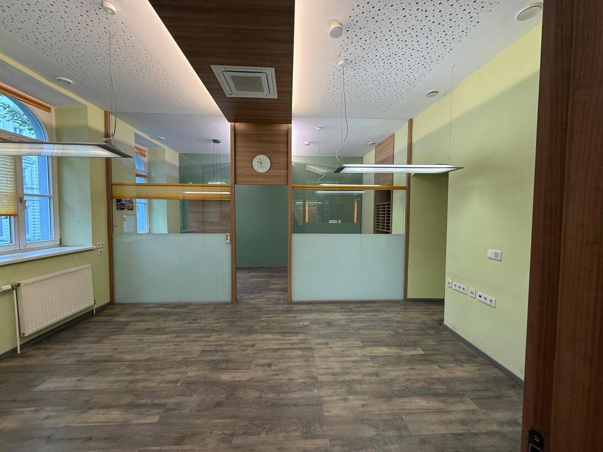 Geräumiges Büro mit Holzoptik-Laminatboden, hellgrünen Wänden und modernen Glasabtrennungen für Arbeitsbereiche.
