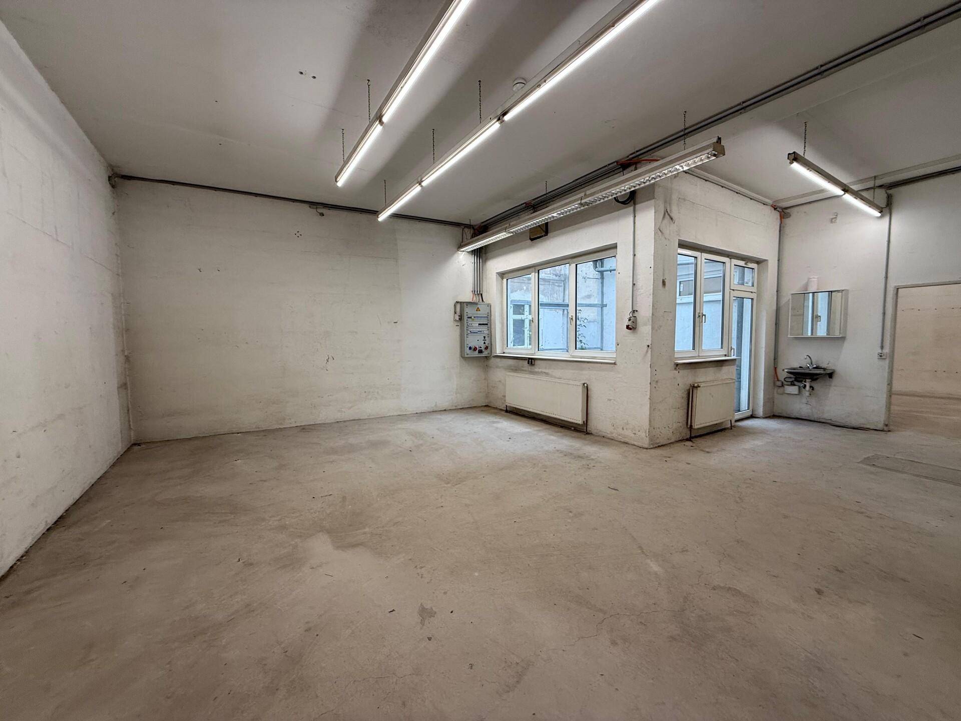 Geräumige Gewerbefläche mit Betonboden, weißen Wänden und großen Fenstern, ideal für Werkstatt oder Lager.