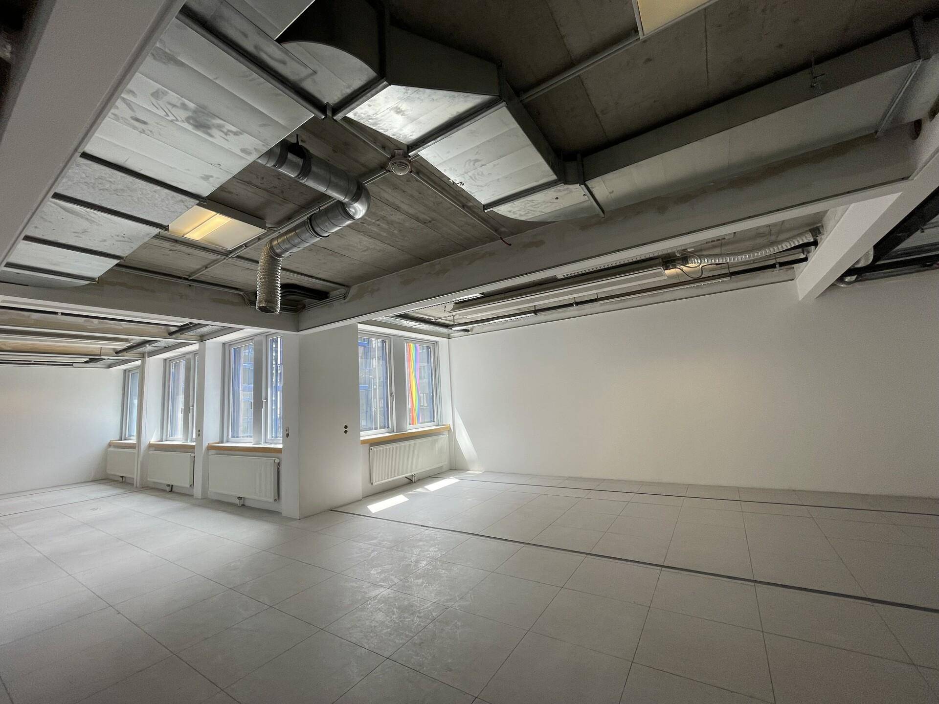 Leerer Büroraum mit Fliesenboden, großen Fenstern und sichtbaren Lüftungsrohren an der Decke.