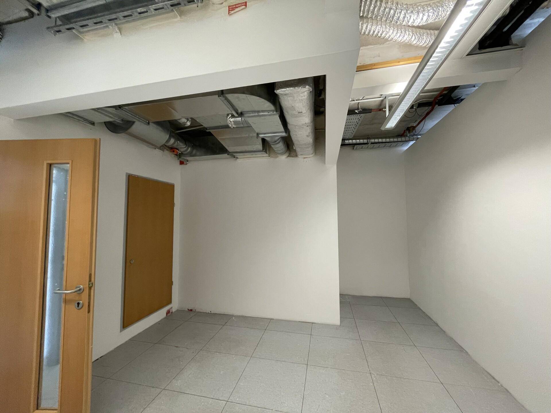 Leerer Raum mit Fliesenboden, weißen Wänden und sichtbaren Lüftungsrohren an der Decke.