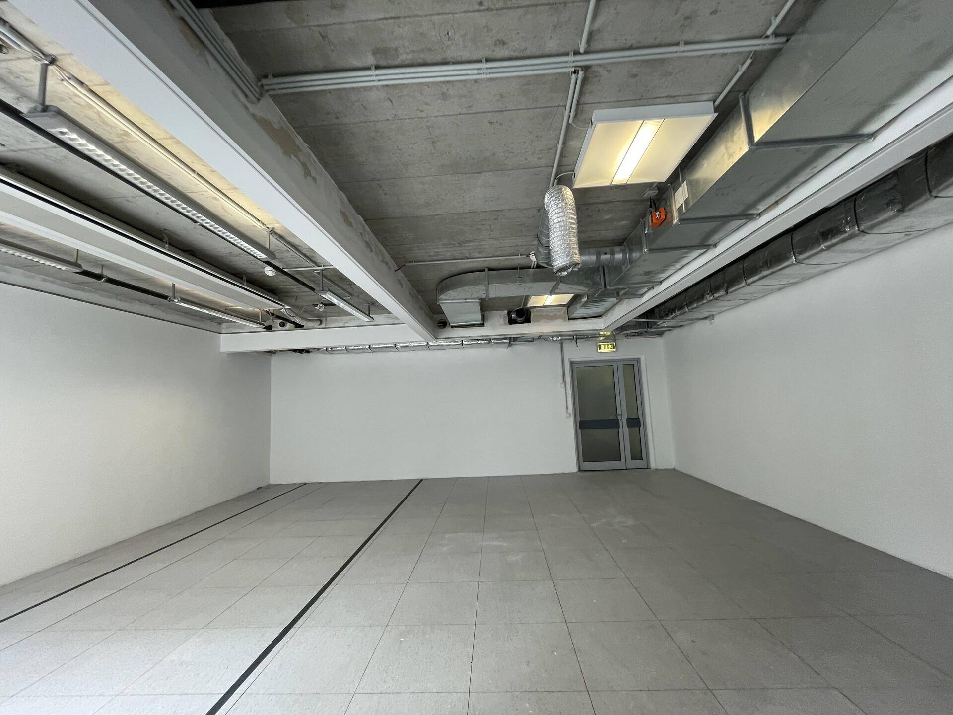 Leerer Büroraum mit Fliesenboden, weißen Wänden und sichtbaren Lüftungsrohren an der Decke.