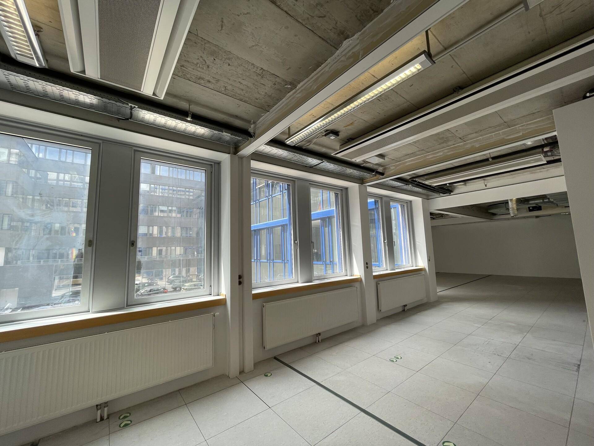 Leerer Büroraum mit Fliesenboden, großen Fenstern und sichtbaren Lüftungsrohren an der Decke.
