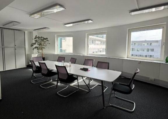 Heller Konferenzraum mit großem weißen Tisch, modernen Stühlen und Fenstern mit Stadtblick.