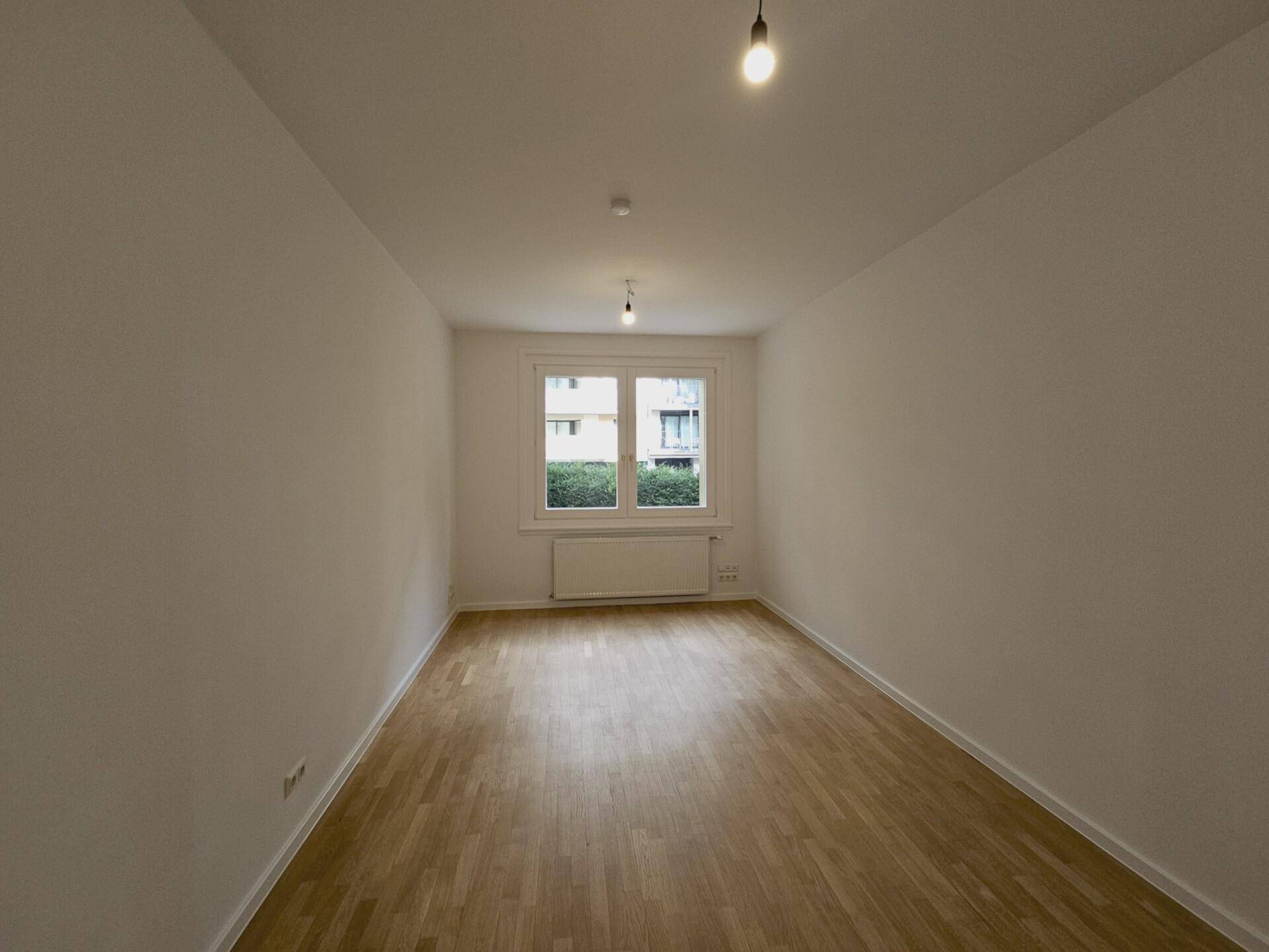 Leeres Schlafzimmer mit hellem Parkettboden und einem Fenster mit Blick auf die Umgebung.