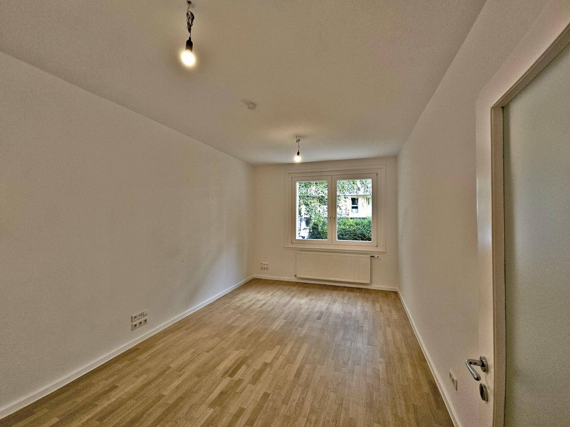 Heller, leerer Raum mit Holzboden und einem Fenster, das Tageslicht hereinlässt.