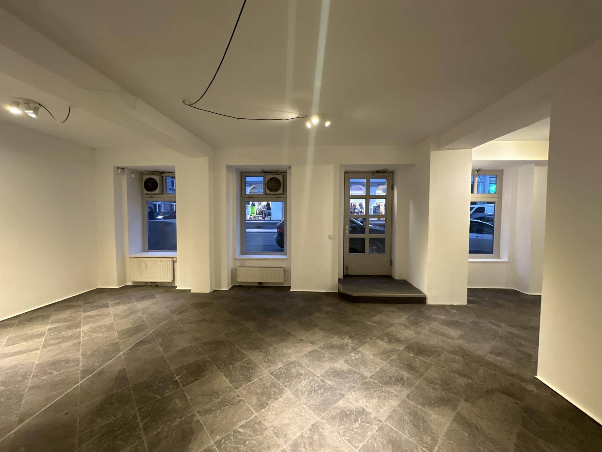 Großer, leerer Geschäftsraum mit dunklem Fliesenboden und mehreren Fenstern, die viel Tageslicht hereinlassen und Klimaanlagen zeigen.