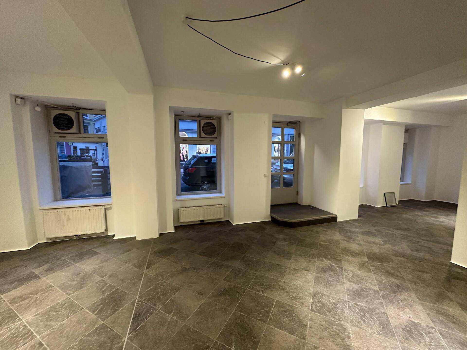 Heller Geschäftsraum mit großen Fenstern zur Straße, dunklem Fliesenboden und sichtbaren Klimaanlagen unter den Fenstern.