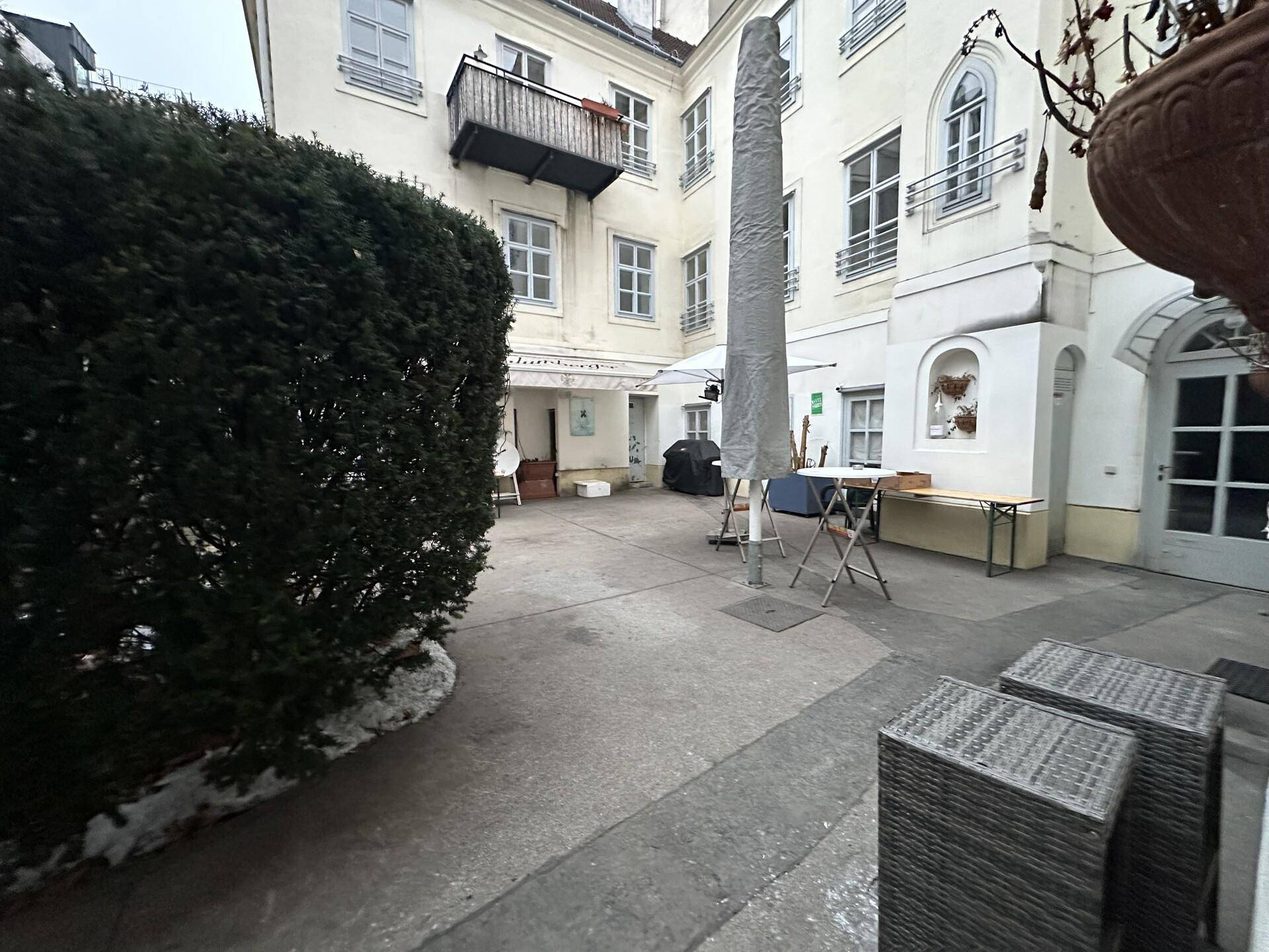 Ruhiger Innenhof eines Altbaus mit Sitzgelegenheiten, einem großen Sonnenschirm und grünen Hecken.