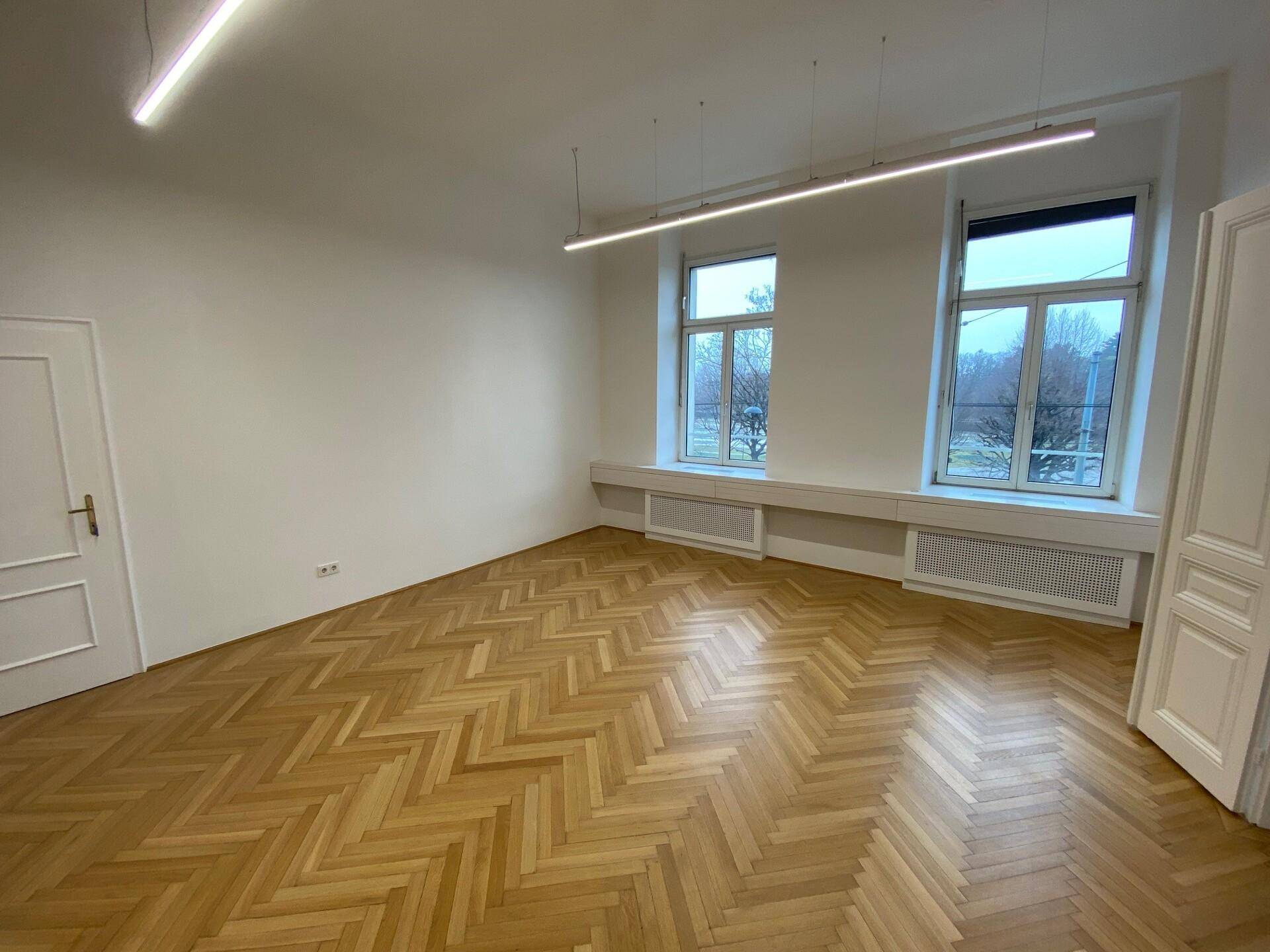 Geräumiger, heller Raum mit Fischgrätparkett, zwei Fenstern, einer klassischen Flügeltür und moderner Beleuchtung.