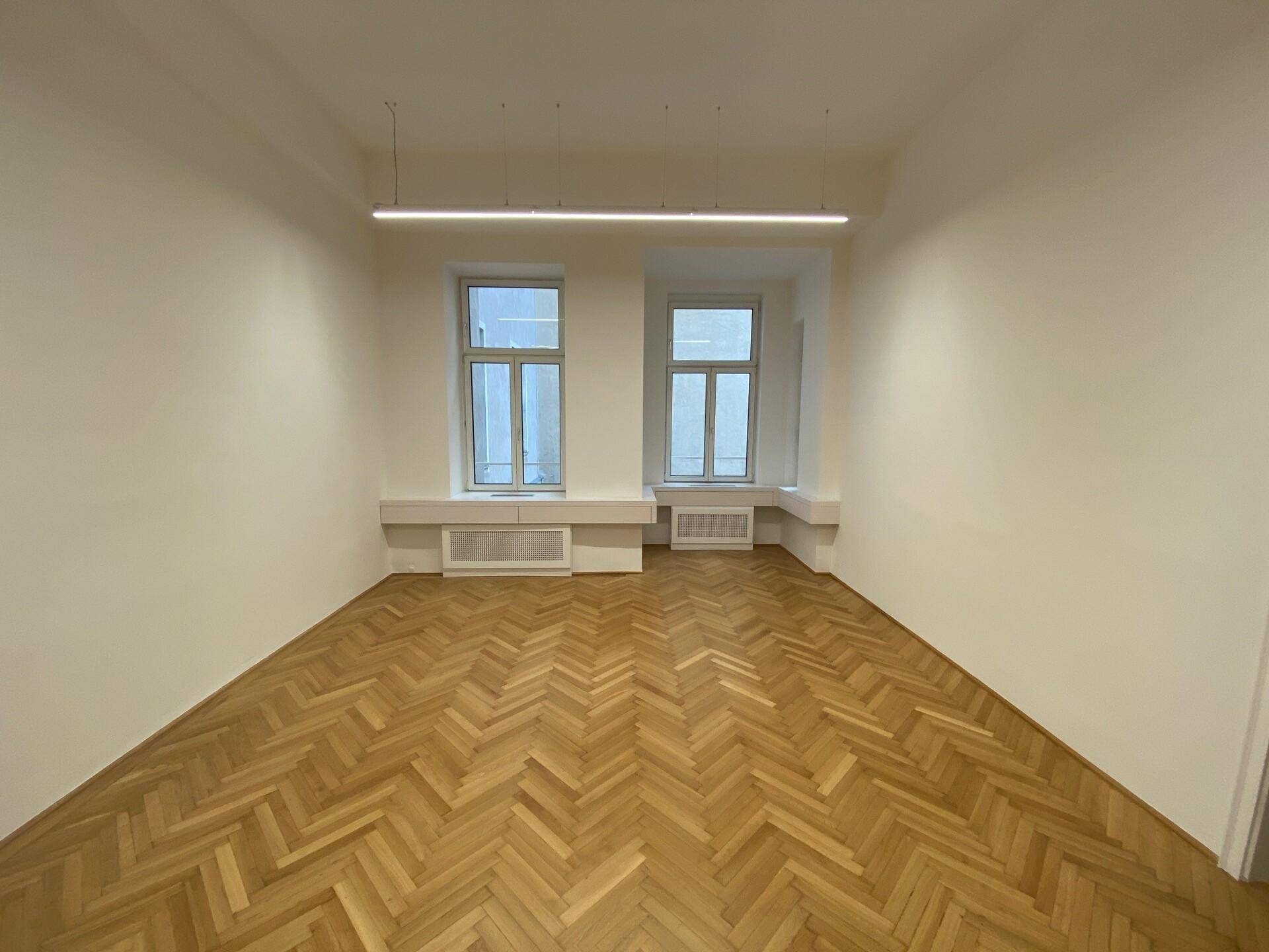 Heller, leerer Raum mit Fischgrätparkett, zwei großen Fenstern und moderner linearer Deckenbeleuchtung.