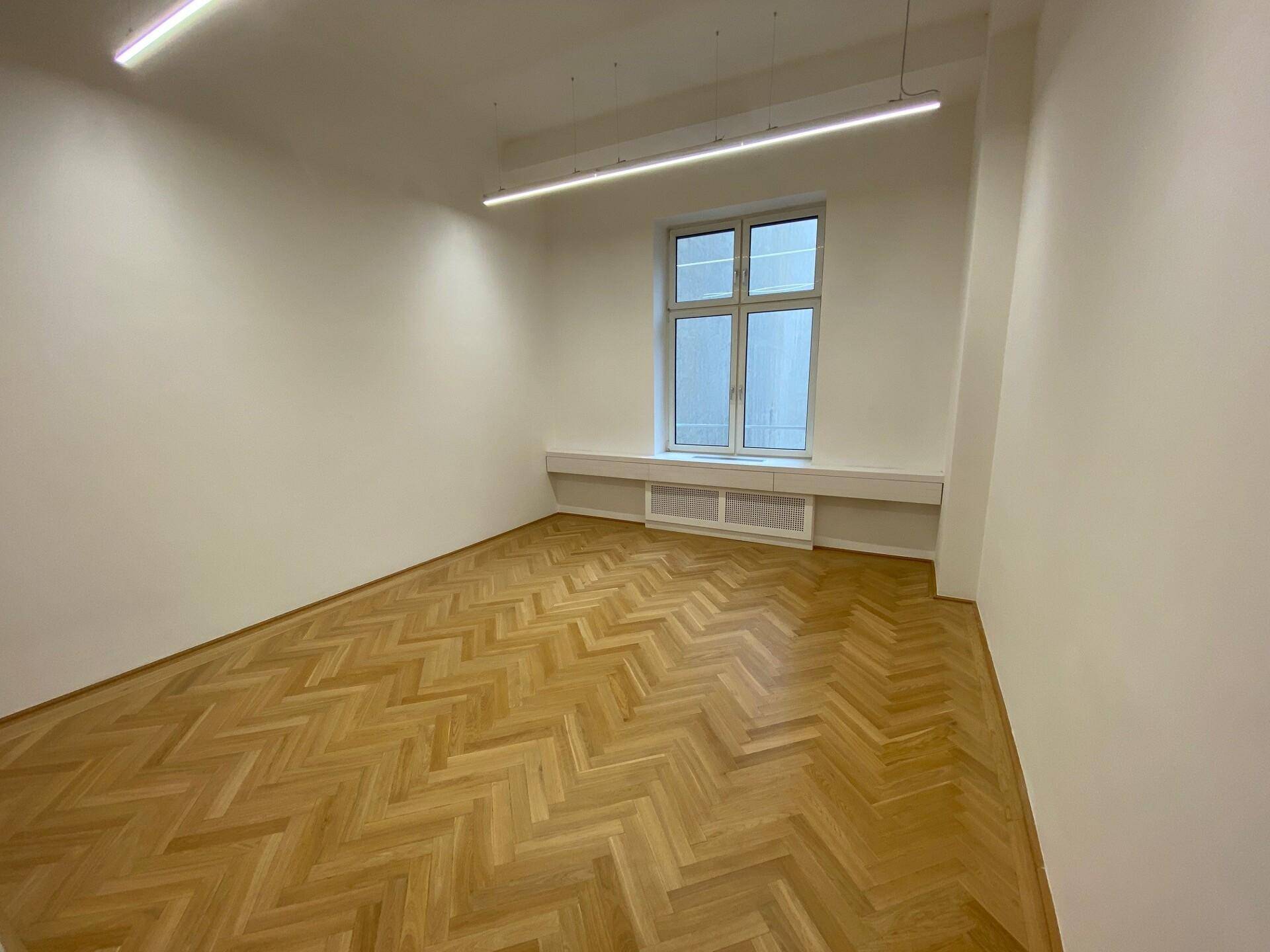 Heller, leerer Raum mit Fischgrätparkett, einem großen Fenster und moderner linearer Deckenbeleuchtung.