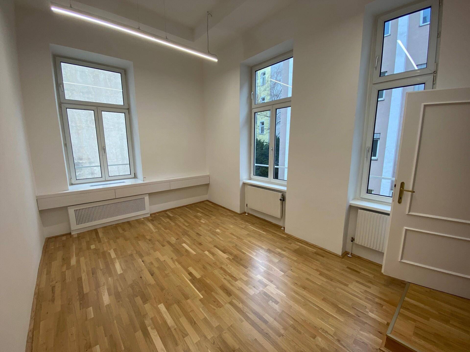 Heller Raum mit Fischgrätparkett, drei Fenstern und einer modernen linearen Deckenleuchte.