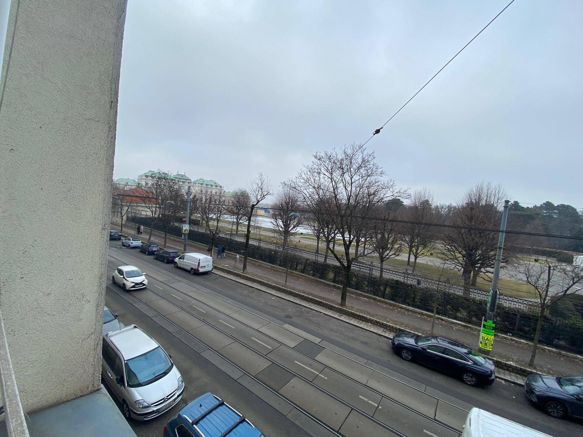 Blick von einem Balkon oder Fenster auf eine belebte Straße mit Park und Bäumen im Hintergrund.
