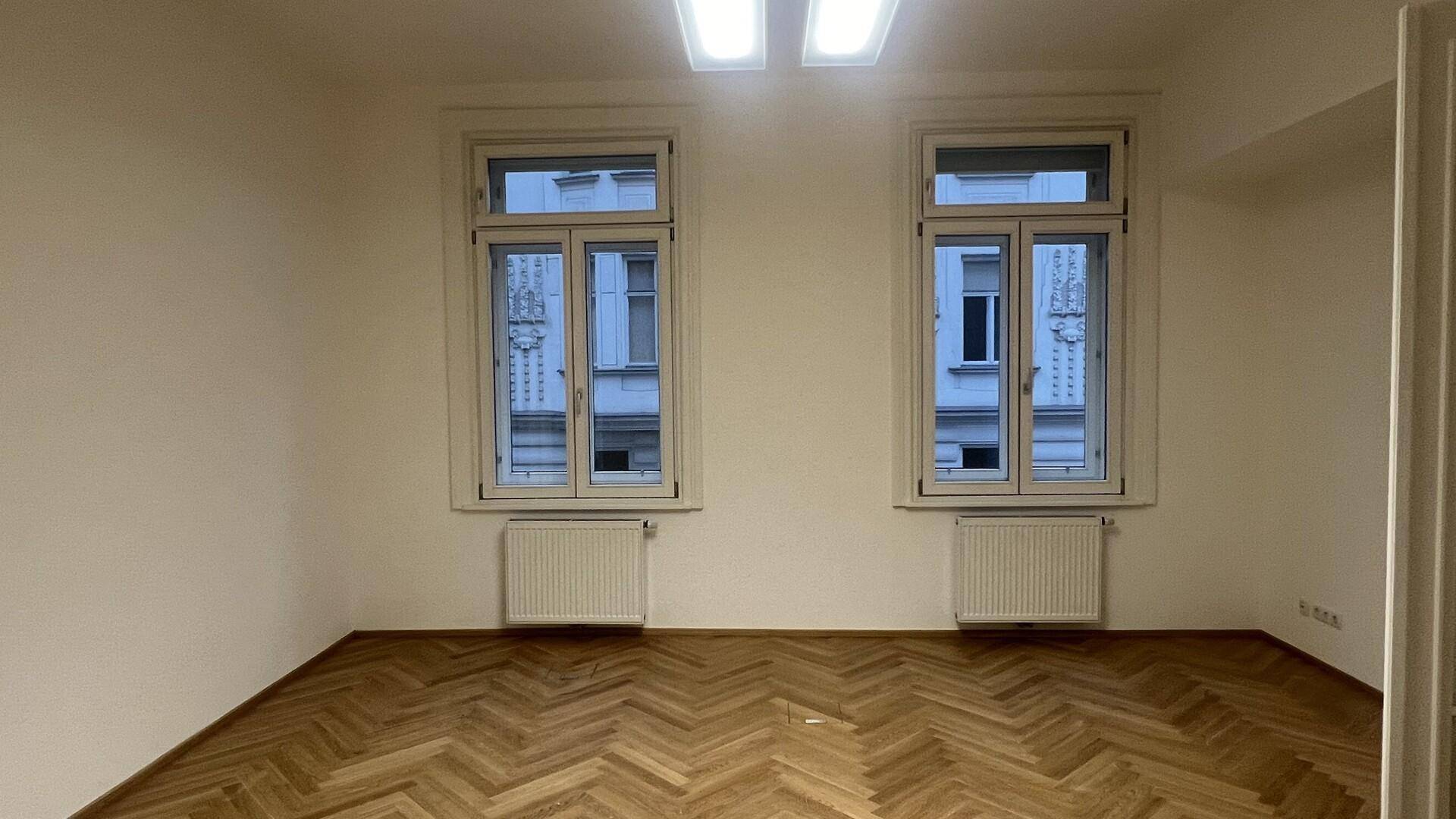 Geräumiger Raum mit zwei hohen Fenstern und elegantem Fischgrätparkett, ideal für verschiedene Nutzungen.