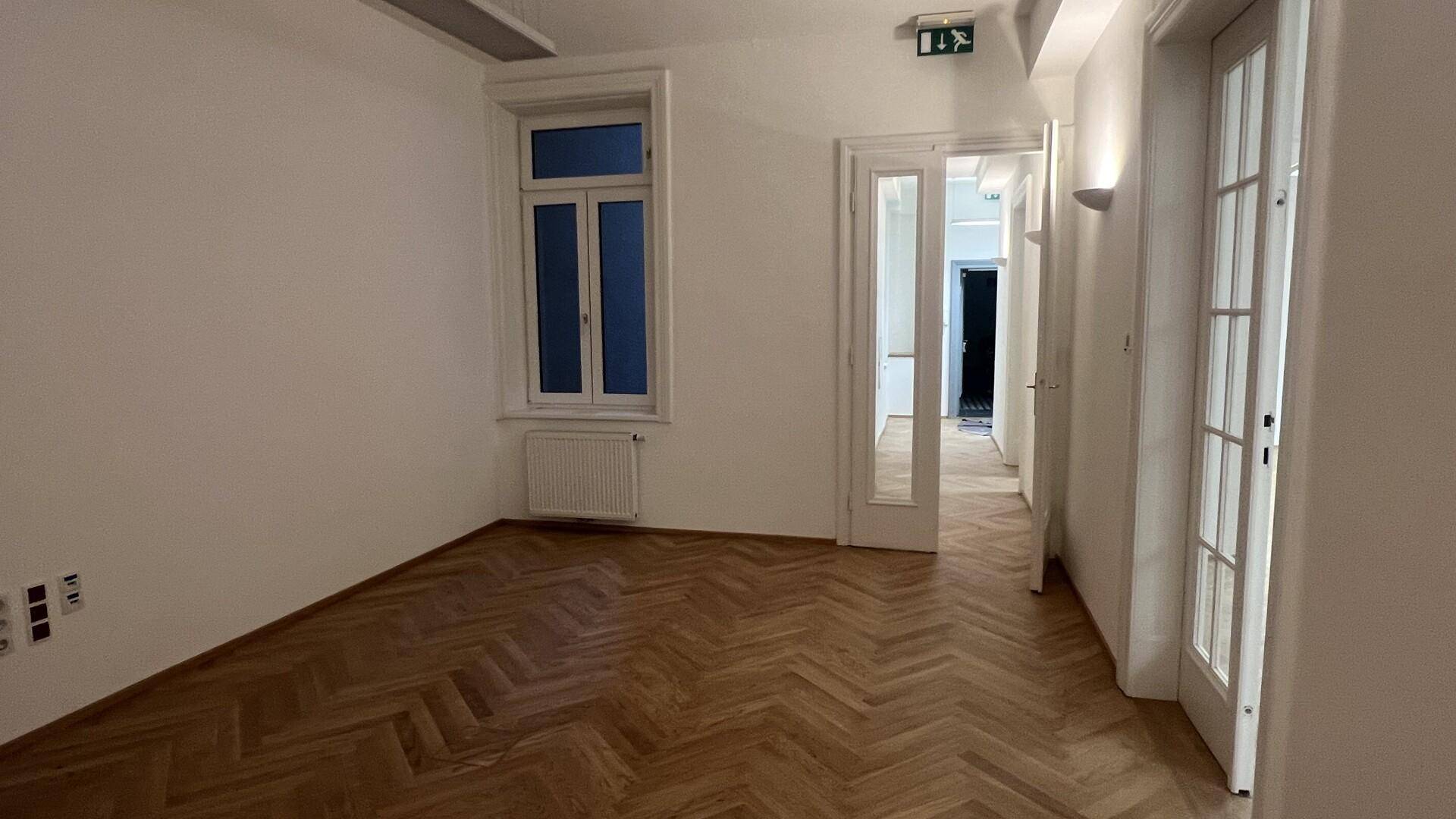 Leerer Raum mit einem Fenster, Fischgrätparkett und einer Tür, die zu einem Flur führt.