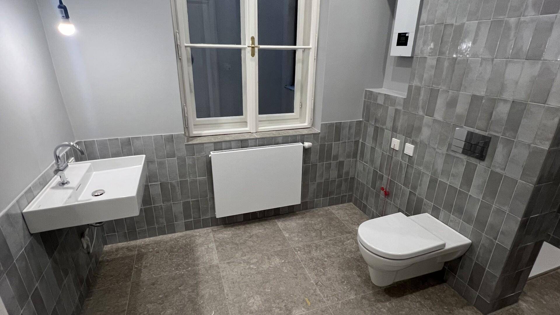 Modernes Badezimmer mit Fenster, Waschbecken und wandhängender Toilette, in Grautönen gehalten.